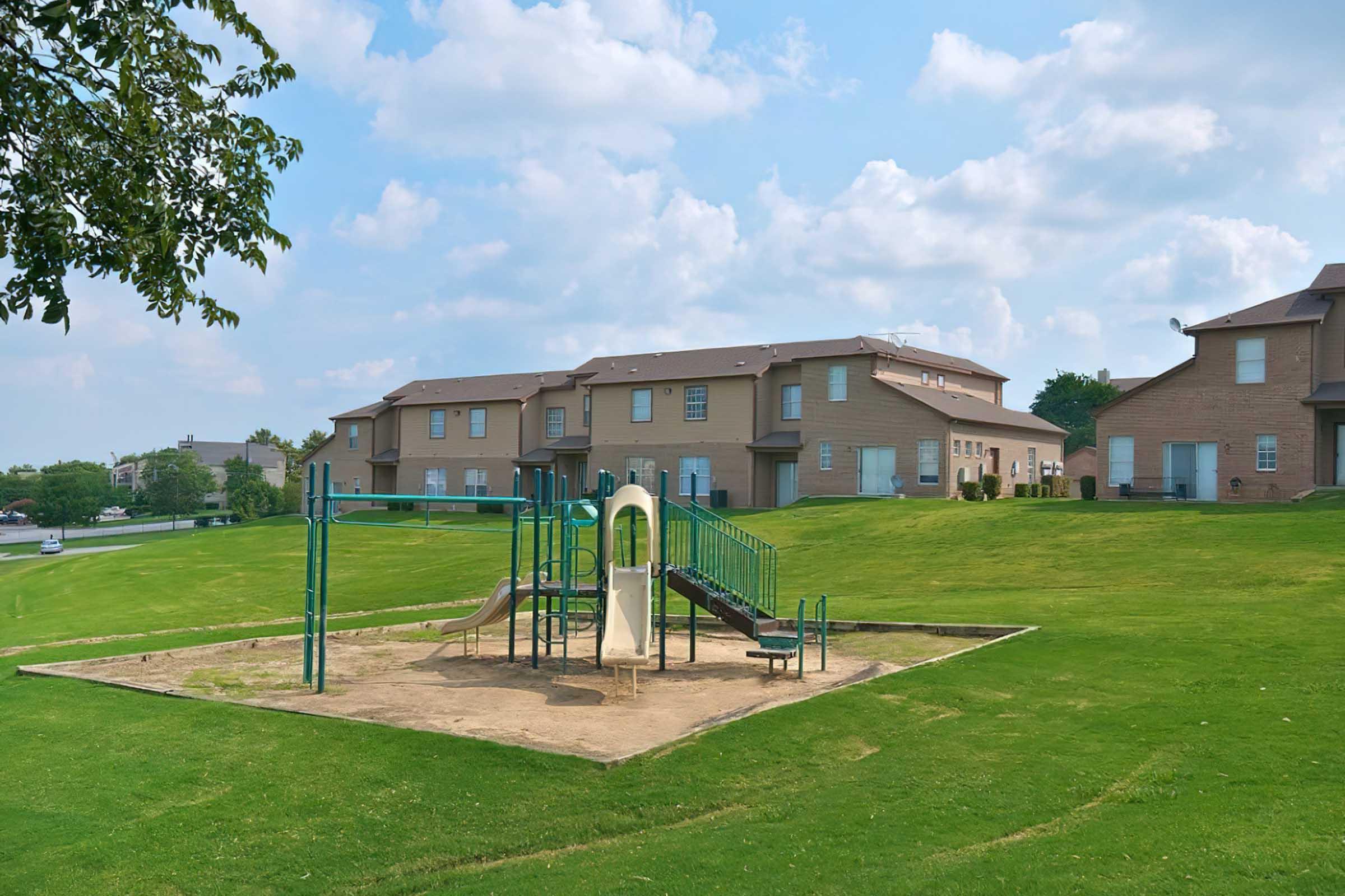 A playground area featuring a slide and climbing structure, situated on a grassy expanse. In the background, there are multi-story brick apartment buildings under a partly cloudy sky. The scene is tranquil and inviting, suitable for families and children.