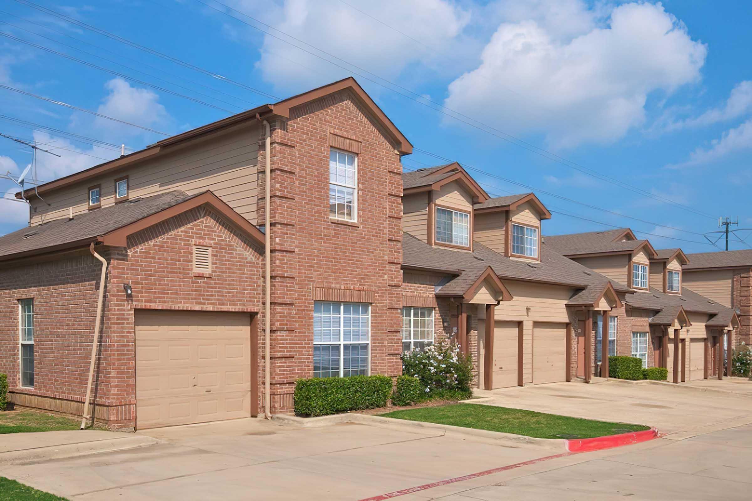 A row of modern brick townhouses with tan accents, featuring attached garages and well-maintained green lawns. The sky is partly cloudy, adding a bright and inviting atmosphere to the suburban neighborhood.