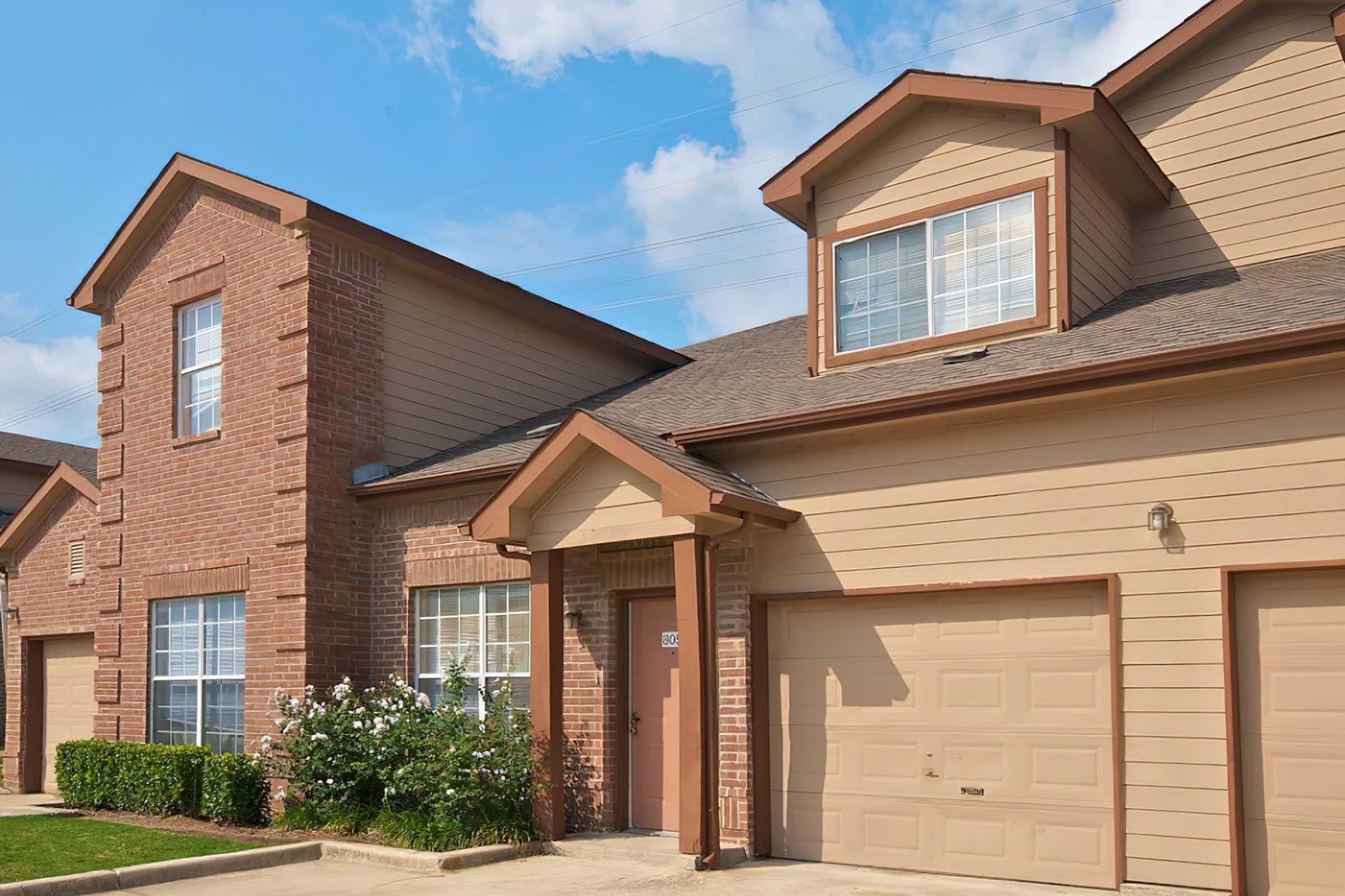 A two-story residential building featuring a combination of brick and wooden siding. The architectural style includes large windows and a gabled roof. The front yard has neatly trimmed greenery, and there are two garage doors visible. The scene is set under a partly cloudy sky.