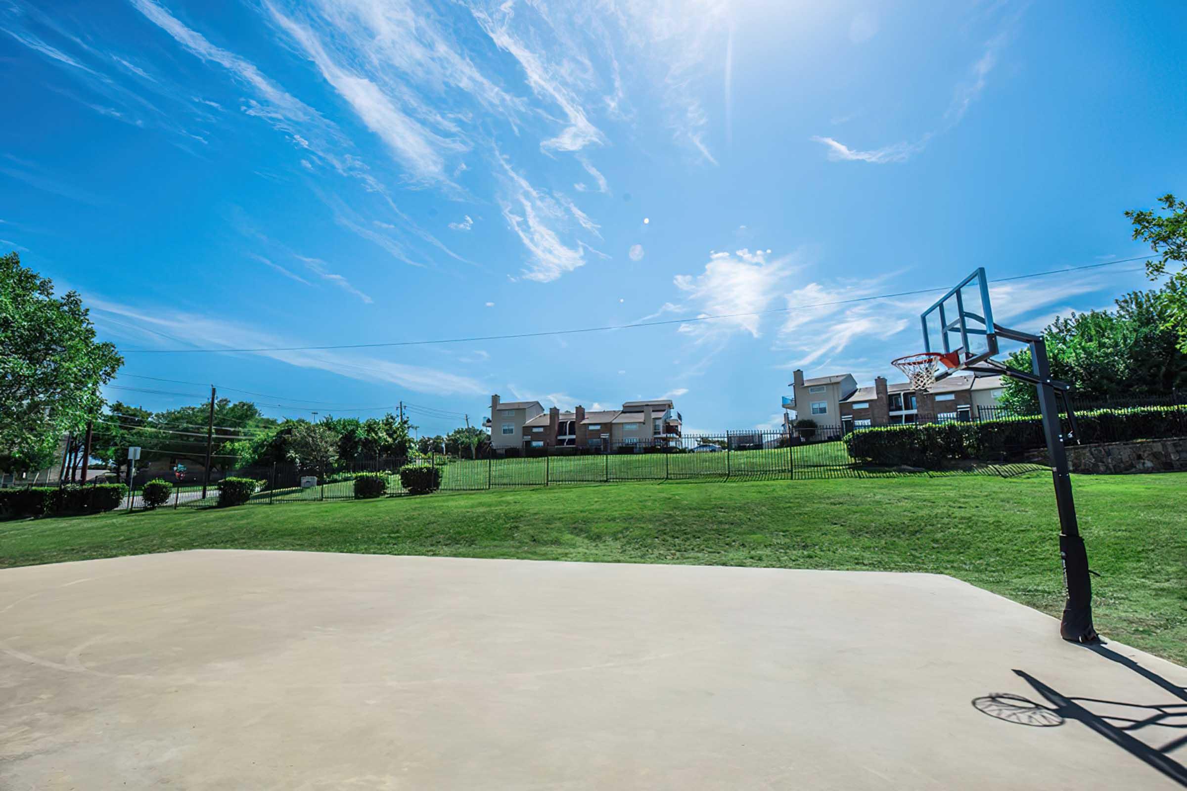 A sunny outdoor basketball court with a clear blue sky and wispy clouds. In the background, there are green grassy areas and several buildings surrounded by trees and shrubs, creating a vibrant community atmosphere.