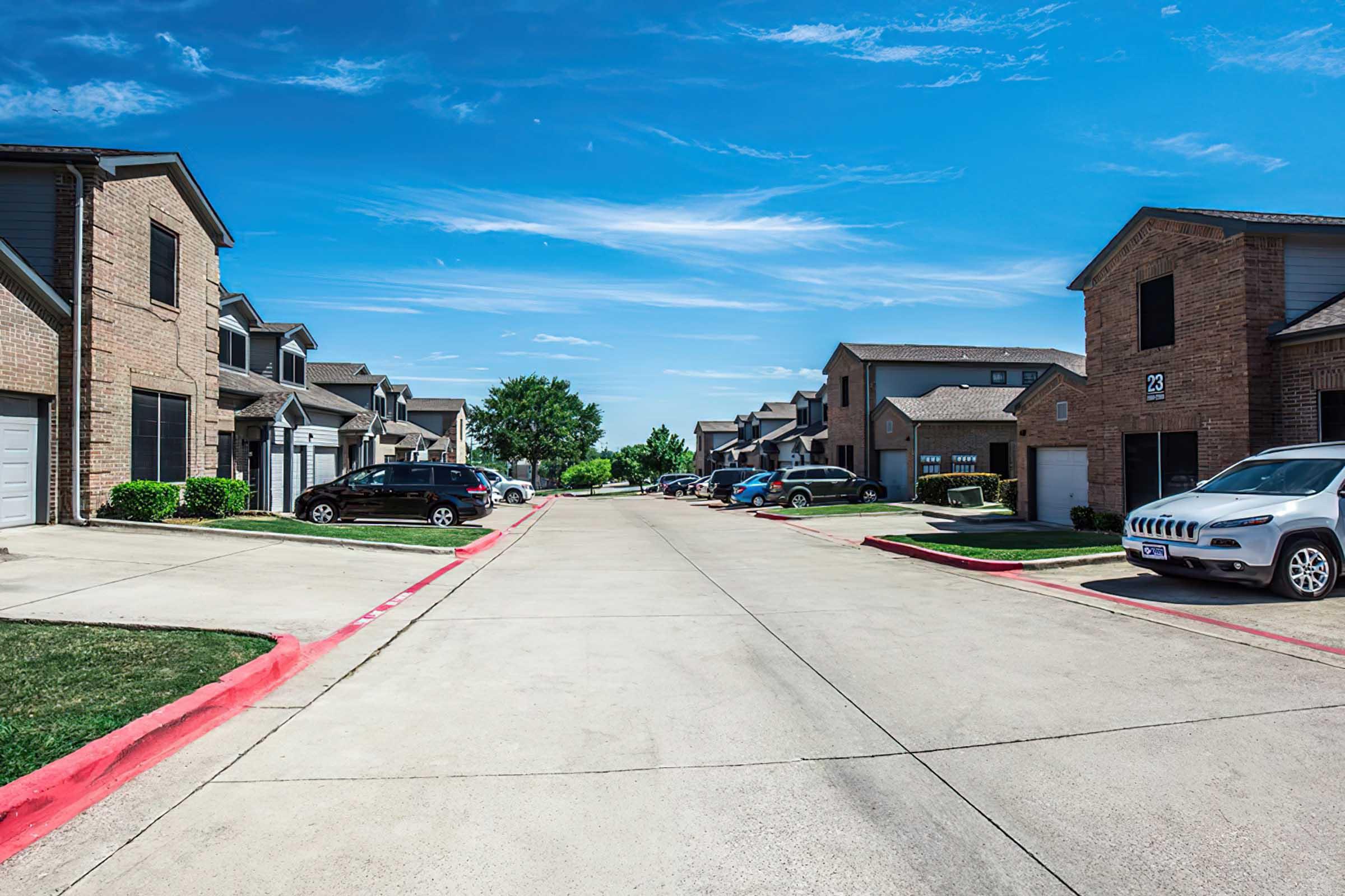 A view of a residential street lined with two-story apartment buildings. The street features parked cars on both sides, and well-maintained lawns with some shrubs. The sky is clear and blue, suggesting a sunny day.