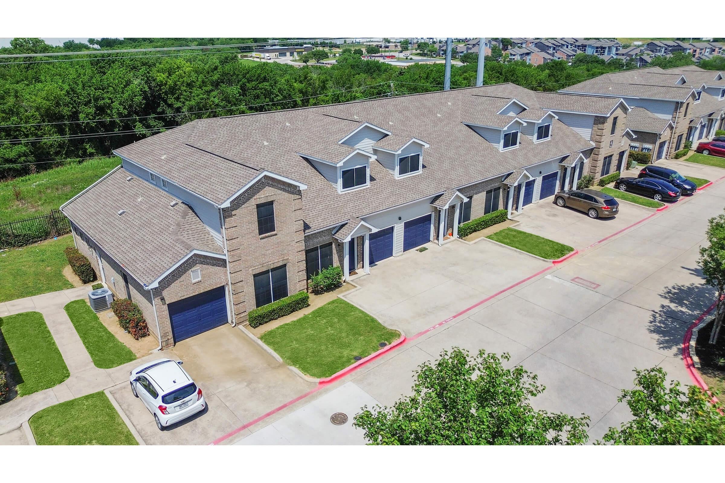 Aerial view of a row of two-story brick townhouses with blue garage doors. There are green lawns in front and a paved parking area with parked cars. The background features trees and additional suburban development. Bright, sunny day with clear skies.
