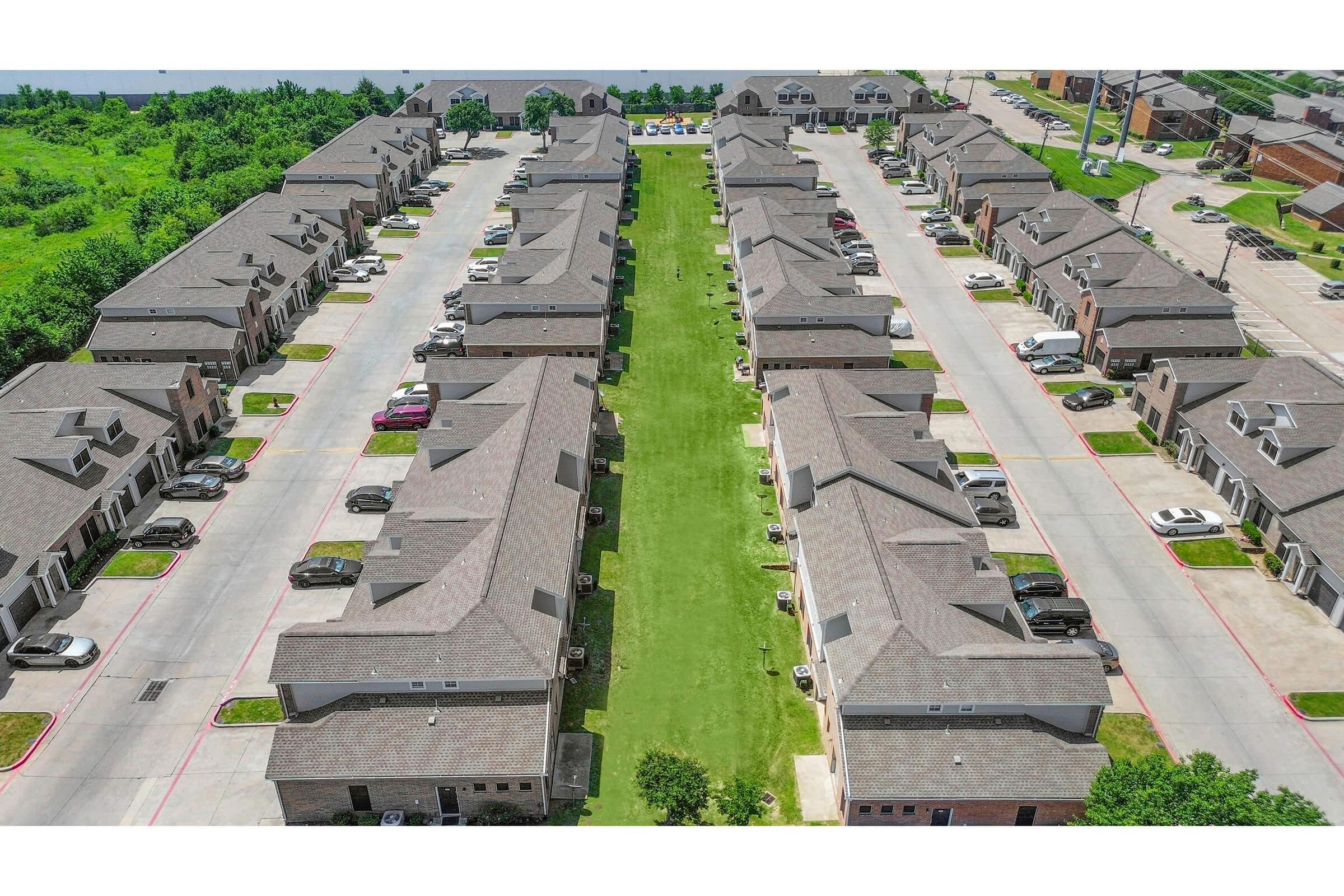 Aerial view of a residential complex featuring multiple rows of attached townhouses. Each unit has a parking area, and a well-maintained green lawn runs between the rows. Surrounding the complex are trees and additional buildings, indicating an urban or suburban setting.