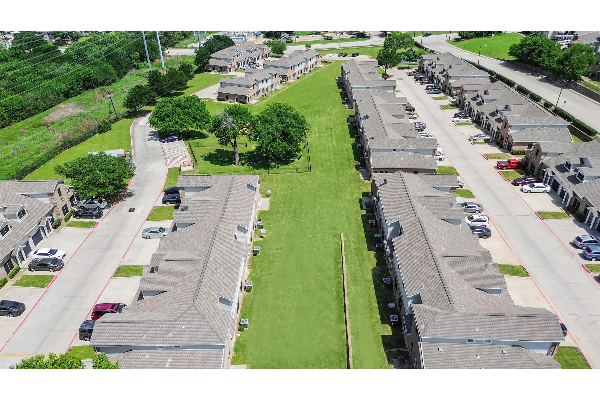 Aerial view of a residential complex featuring multiple two-story buildings arranged along pathways, with green lawns in between. Several parked cars are visible along the driveways, and trees are scattered throughout the area, providing shade. The surrounding landscape includes roads and open fields.
