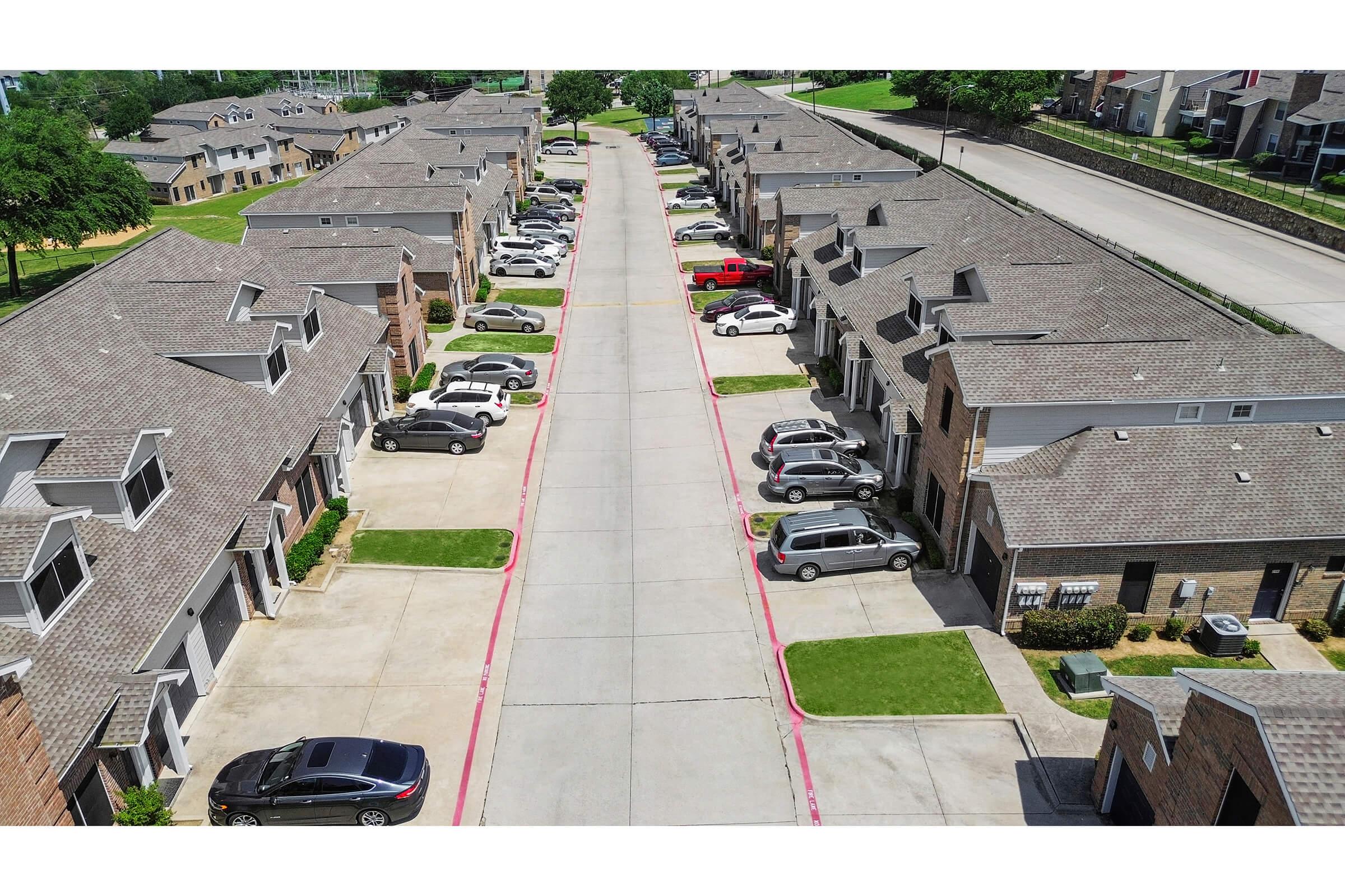 Aerial view of a residential complex featuring a tree-lined street lined with parked cars on both sides. The buildings consist of two-story townhouses with sloped roofs. The area is well-maintained with grassy lawns and an overall tidy appearance.