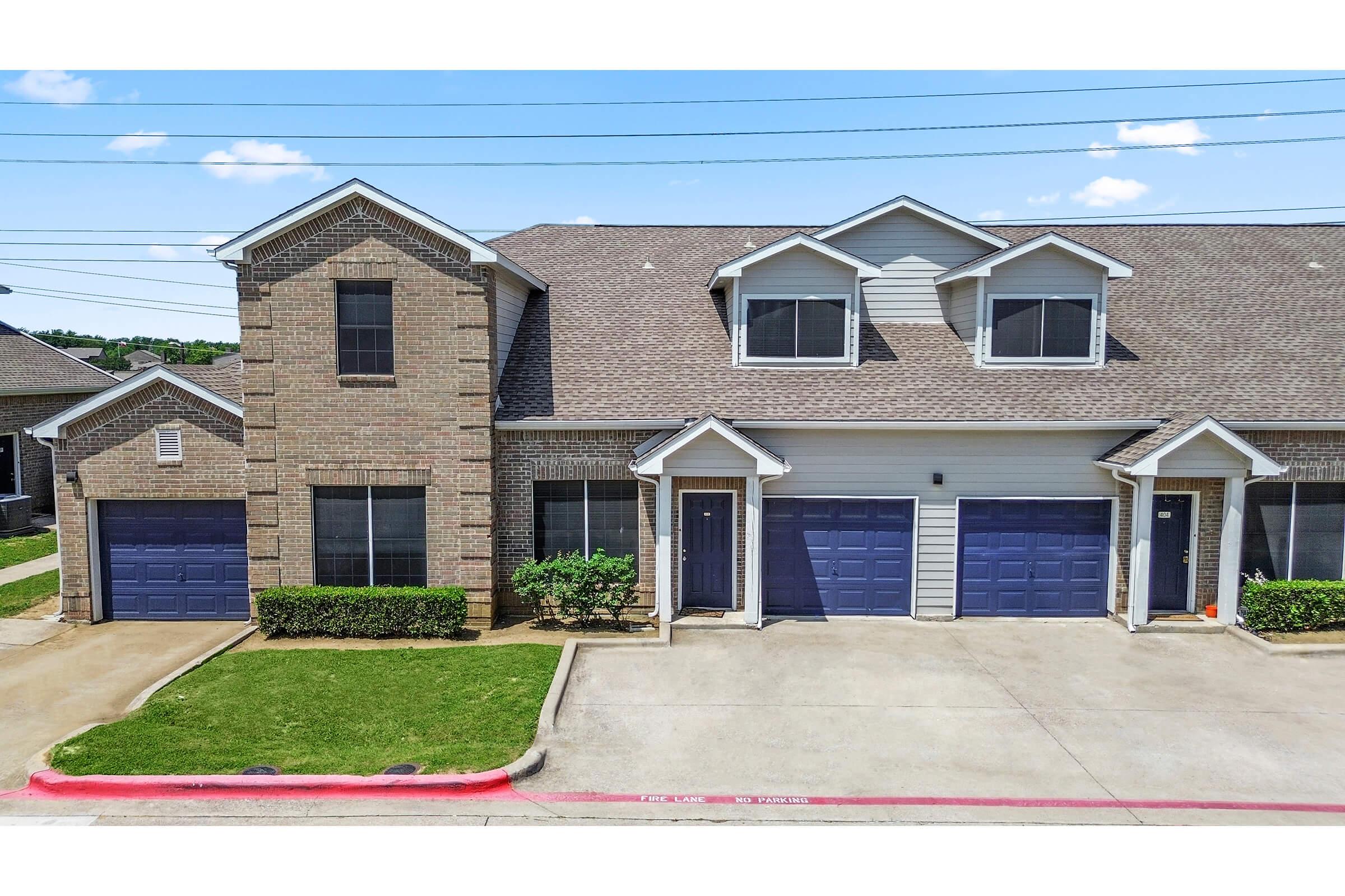 Two-story townhouse with a brick facade, featuring three front doors and matching blue garage doors. The property has manicured shrubs along the walkway and is situated in a sunny area with clear blue skies above. Power lines are visible in the background.