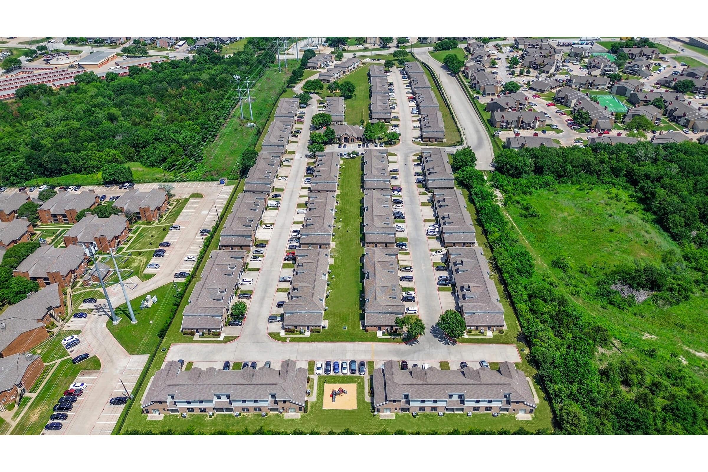 Aerial view of a residential complex featuring multiple buildings arranged in rows, with parking spaces visible in front of each unit. Surrounding greenery and power lines are also present, alongside additional residential areas and roads in the background. The layout highlights organized housing with outdoor spaces.
