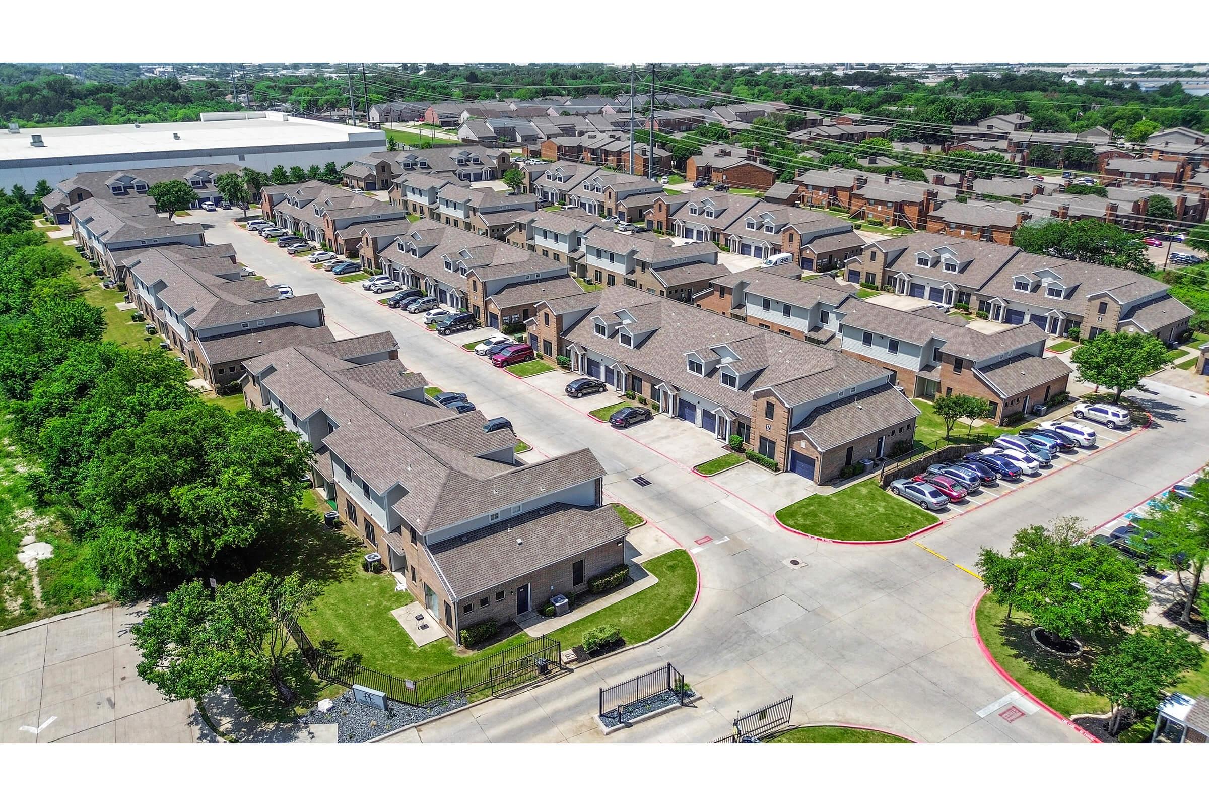 Aerial view of a residential neighborhood featuring multiple two-story buildings with brown roofs. The layout includes several streets lined with parked cars and green landscapes, including lawns and trees, creating a suburban environment.
