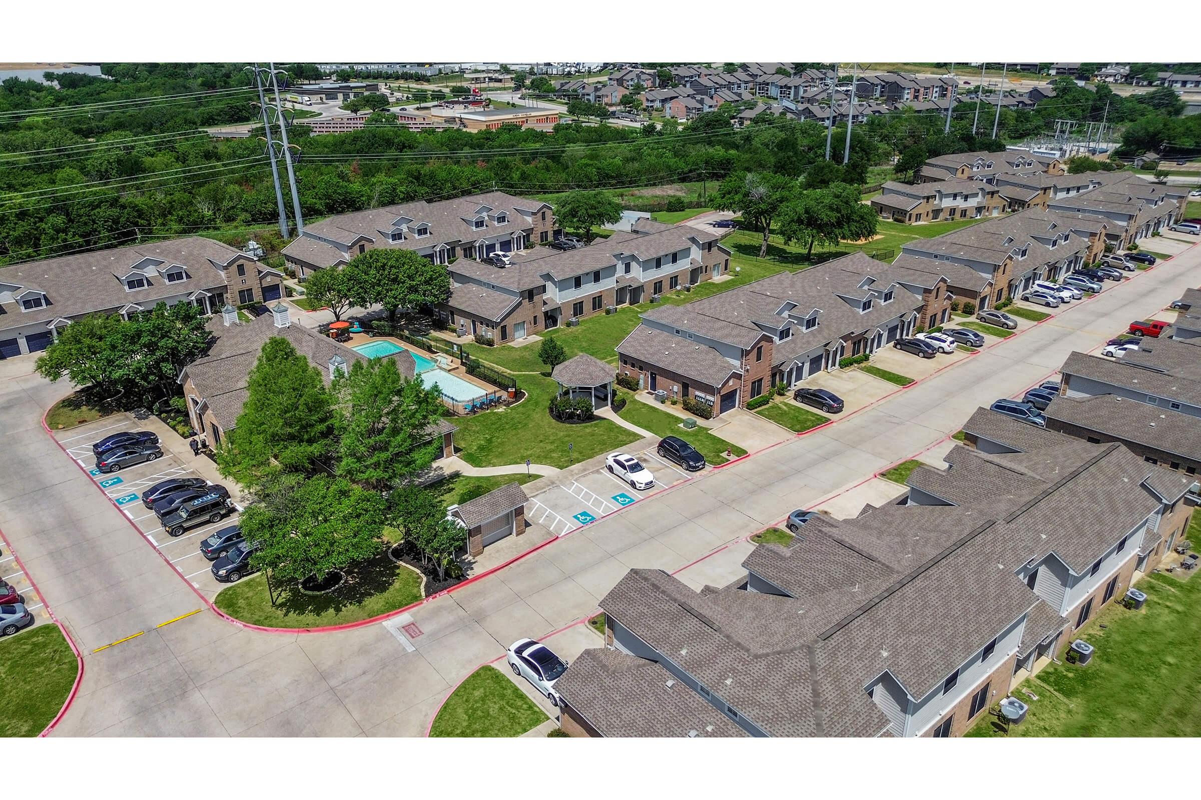 Aerial view of a residential neighborhood featuring multiple multi-unit buildings with a swimming pool and gazebo. The area is landscaped with trees and greenery, and several parked cars line the streets. Power lines are visible in the background, along with more buildings in the distance.