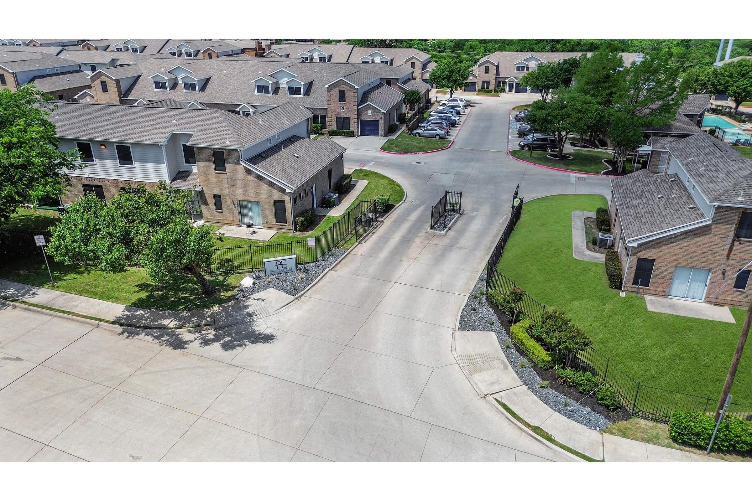 Aerial view of a residential neighborhood featuring a paved road leading into a gated community. Surrounding buildings are mostly two-story brick apartments with pitched roofs. Lush green lawns and trees line the pathways, while parked cars are visible along the sides of the road. Clear blue sky overhead.