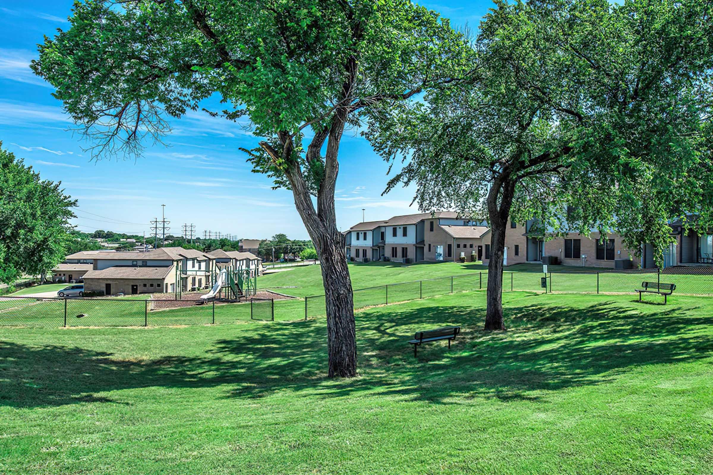 A serene landscape featuring green grass and two large trees. In the background, there are modern buildings with multiple stories, set against a clear blue sky. There are benches scattered throughout the area, and children’s playground equipment can be seen on the left side. The scene conveys a peaceful environment suitable for relaxation and play.