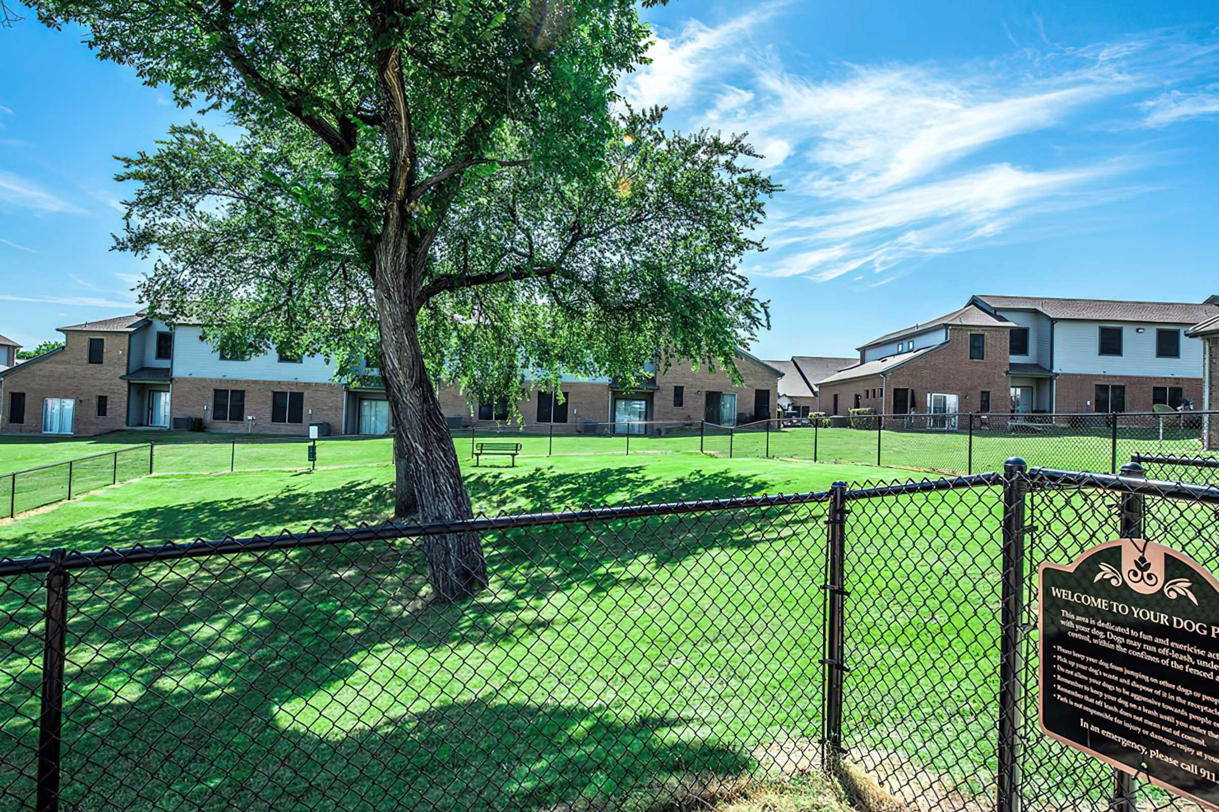 View of a fenced dog park featuring manicured green grass and shade trees. In the background, modern brick buildings are visible, providing a residential area. A sign at the foreground welcomes visitors to the dog park, indicating it's a designated space for dogs to play and socialize.
