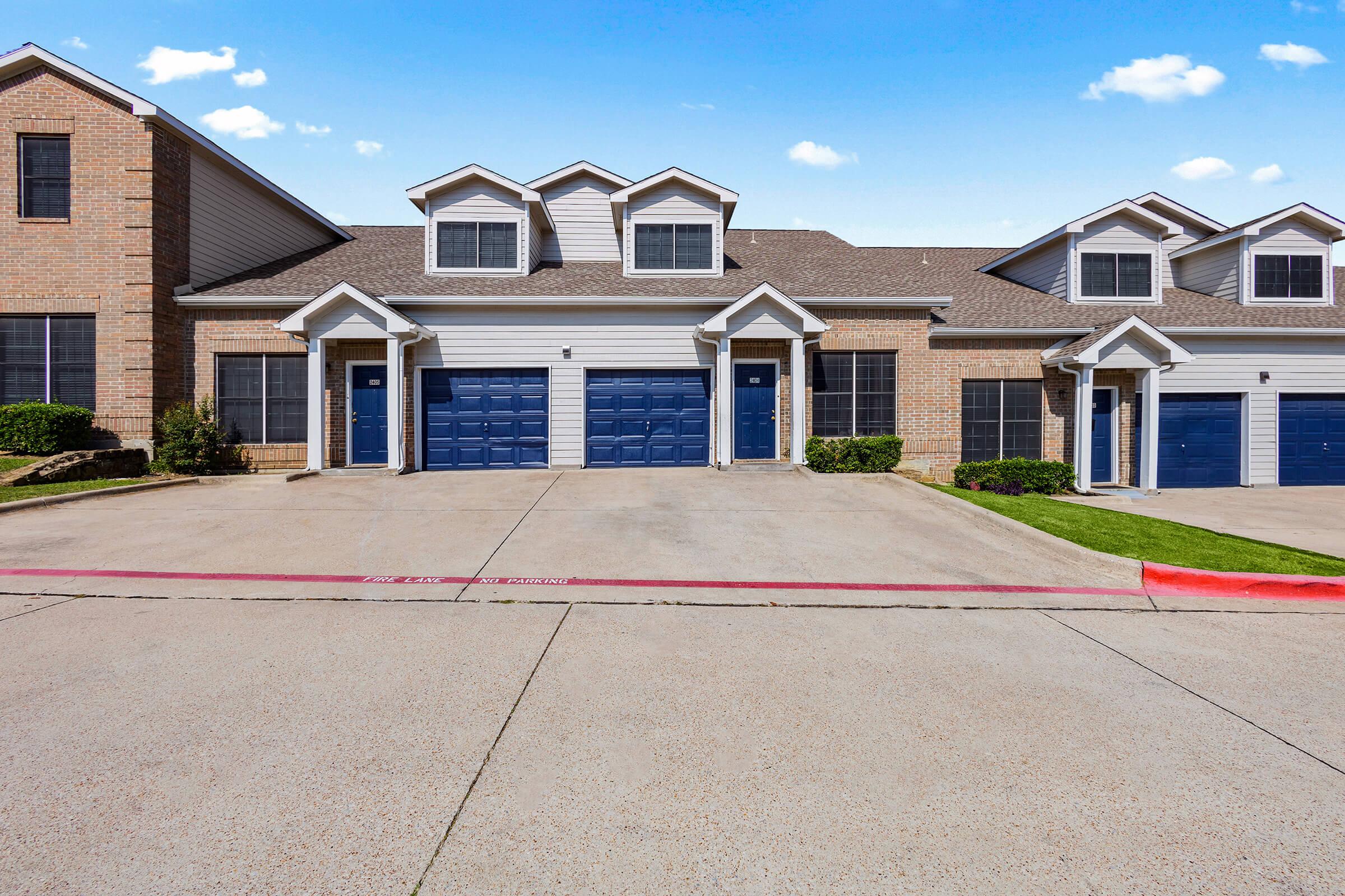 Row of suburban townhouses with beige and gray siding. Each unit features a peaked roof and a blue garage door. The driveway is paved with a red curb at the front, and there are manicured bushes alongside the buildings. The sky above is clear with a few fluffy clouds.