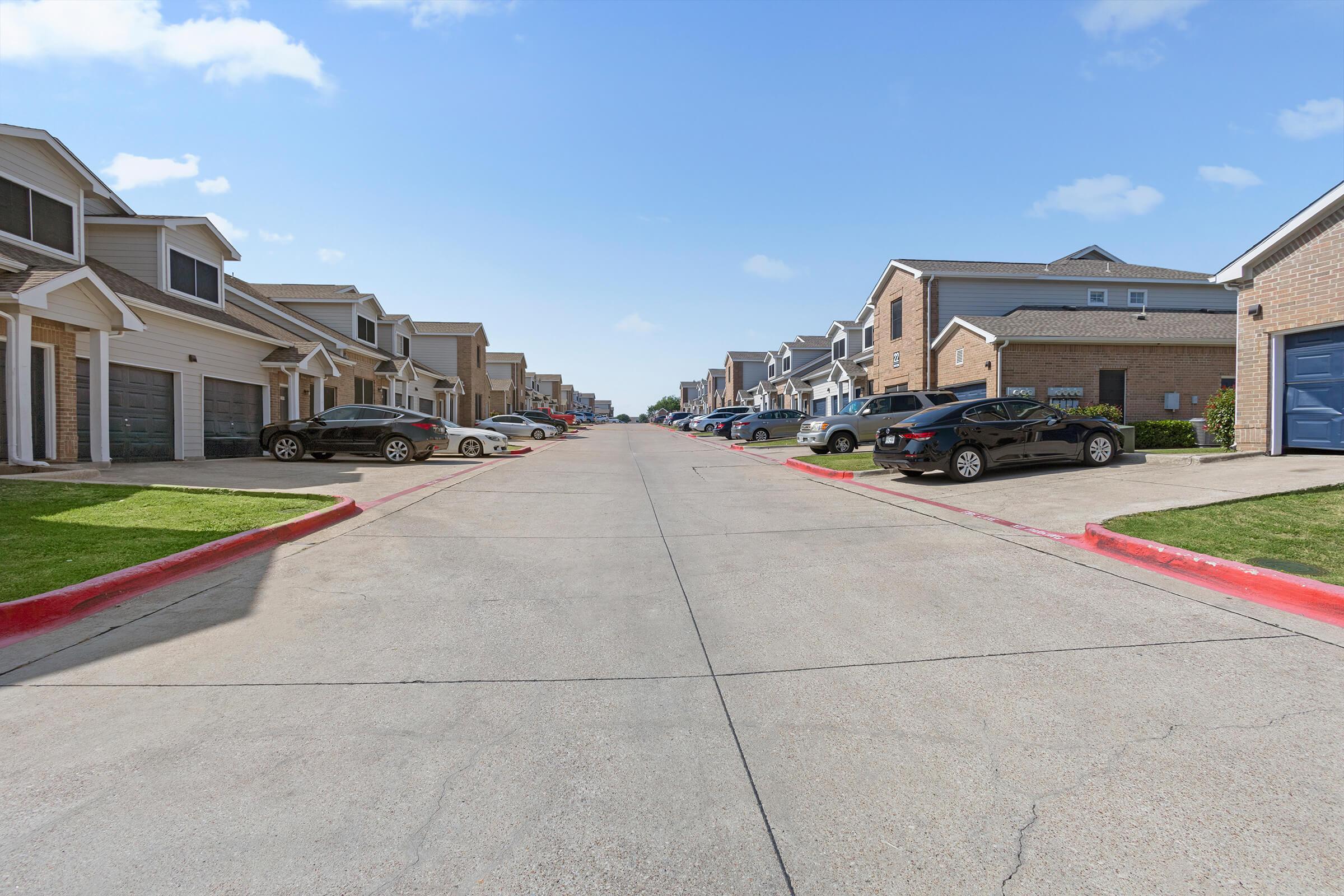 A quiet suburban street lined with townhouses on both sides. Several parked cars are visible along the curbs, and the road is paved with a clear blue sky overhead. The scene conveys a peaceful residential atmosphere.