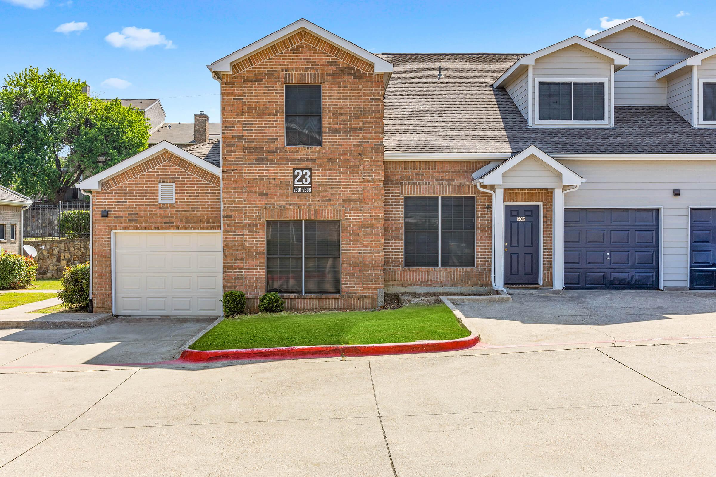 A brick duplex with a number 23 displayed prominently. It features a single-car garage on the left, a dark front door, and multiple windows. The front yard has manicured grass, and the driveway is paved. The setting is sunny, with a clear blue sky and greenery in the background.