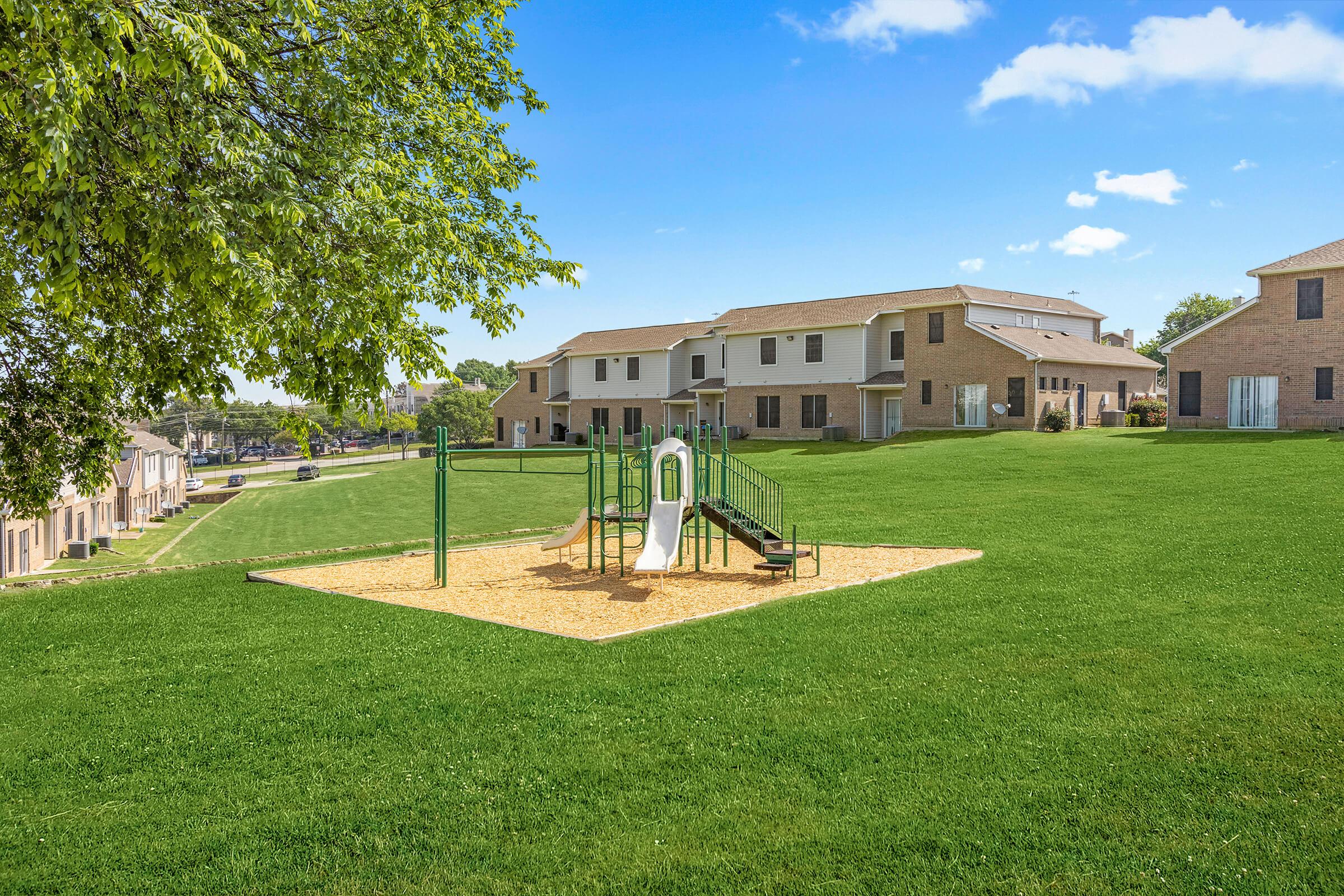 A playground featuring a slide and swings on a grassy area, surrounded by residential buildings under a blue sky with a few clouds. The scene conveys a peaceful, family-friendly environment with ample outdoor space.