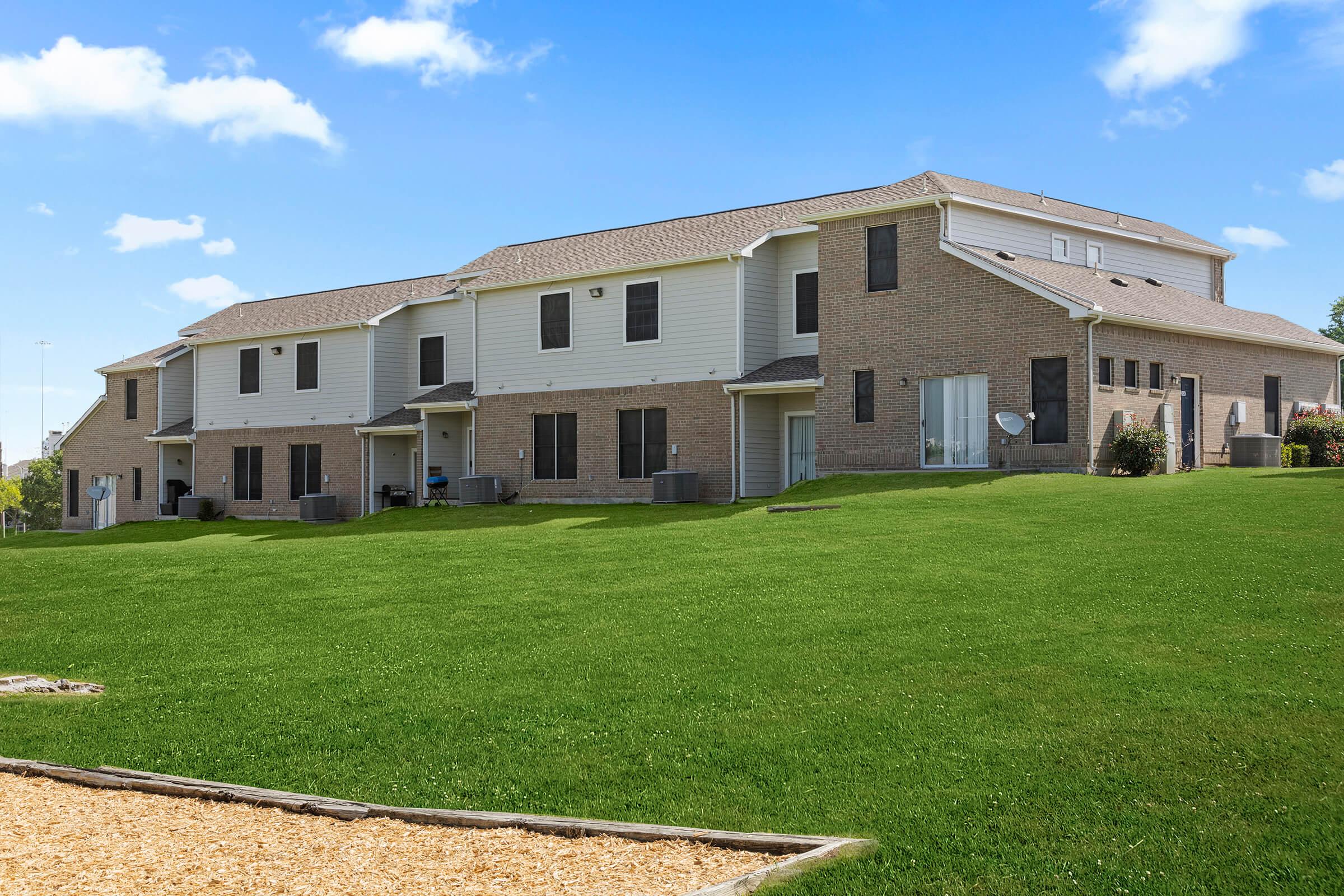 A view of a two-story residential building with a combination of brick and siding exteriors, surrounded by a well-maintained green lawn. The building features multiple windows and doors, with air conditioning units visible outside. A playground area with wood chips is in the foreground under a clear blue sky.