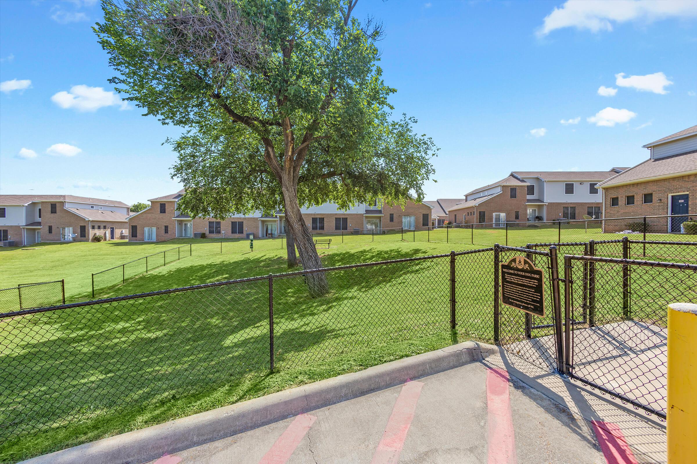 A well-maintained residential area featuring a grassy courtyard surrounded by several two-story brick buildings. A large tree provides shade in the center, and a fenced perimeter encloses the space. The sky is clear with a few clouds, creating a bright and inviting atmosphere.