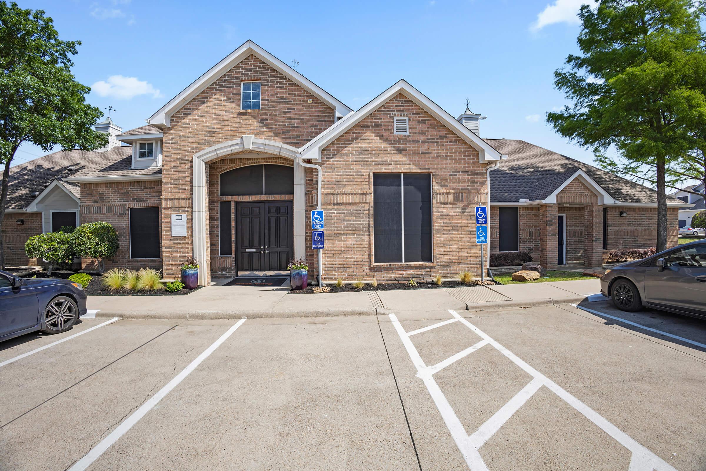 A brick building featuring large windows, a sloped roof, and a welcoming entrance. The surrounding area includes landscaped greenery and parking spaces. Accessible signage is visible near the entrance. The setting appears to be well-maintained and inviting.
