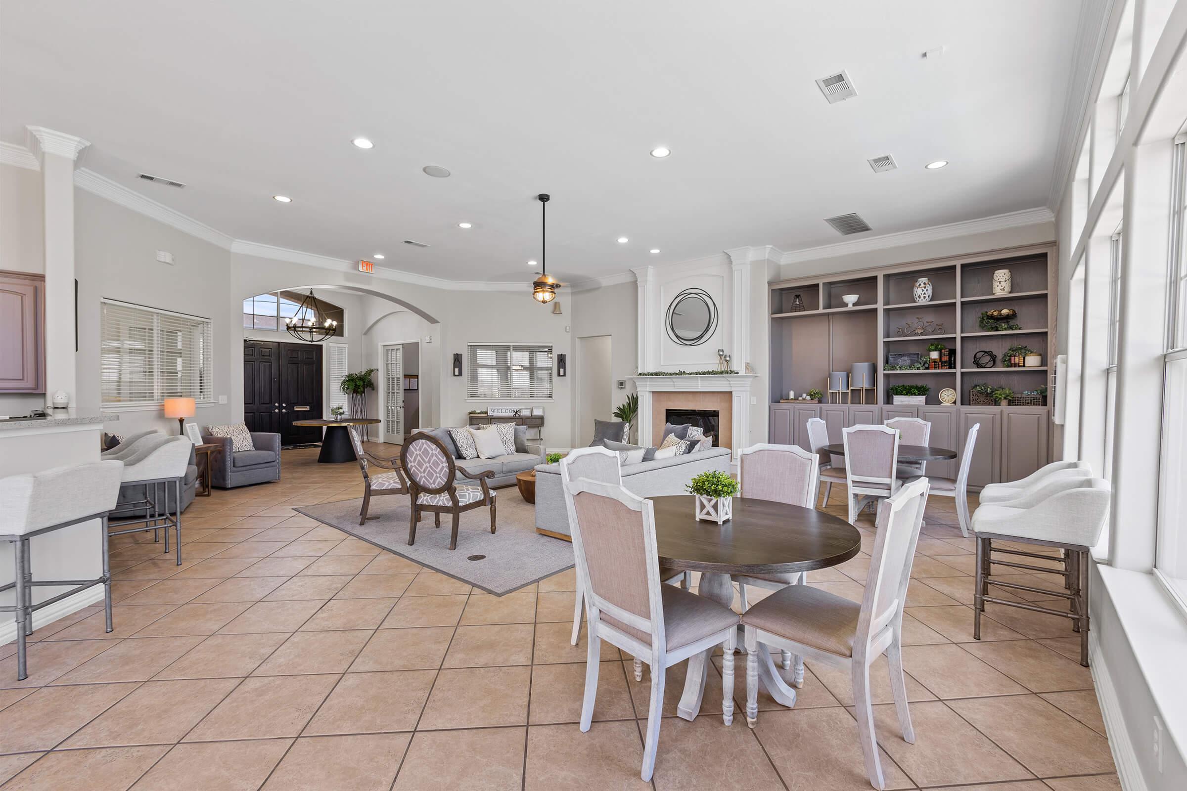 A spacious lounge with a cozy atmosphere featuring a fireplace, comfortable seating, and a dining area. The room has large windows providing natural light, and decor includes plants and stylish furniture. The layout encourages social interaction in a relaxed setting.