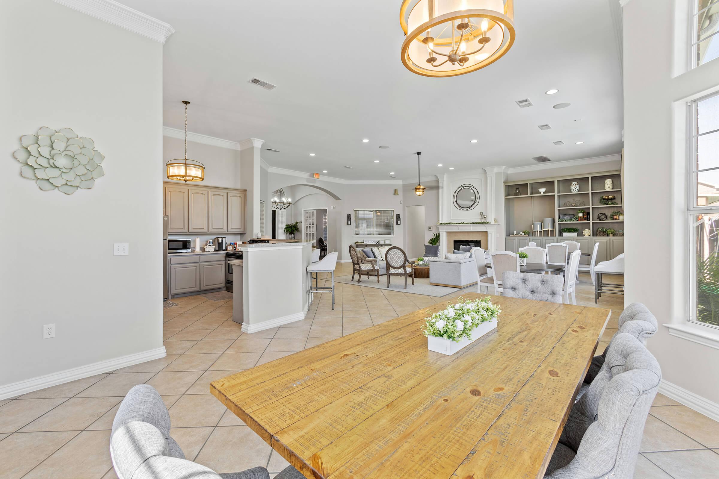 A spacious, modern kitchen and dining area with a large wooden table surrounded by gray upholstered chairs. The kitchen features light cabinets, stainless steel appliances, and an island. Large windows allow natural light, and there’s a cozy living space with a fireplace and elegant decor.