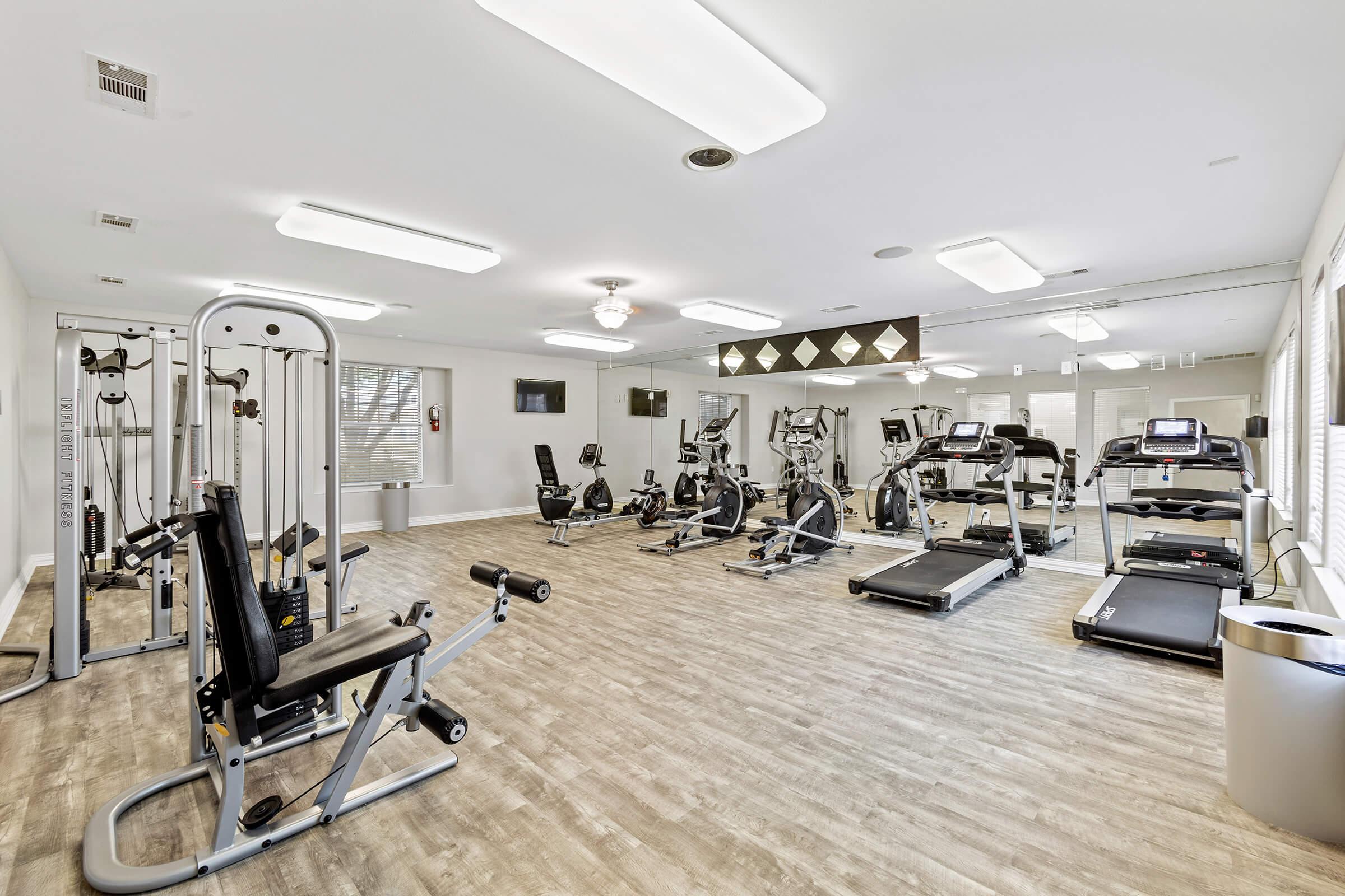 A modern gym interior featuring various fitness equipment, including weight machines, treadmills, and stationary bikes. The space is well-lit with a clean design, hardwood floors, and large windows. A television is mounted on the wall for entertainment during workouts.