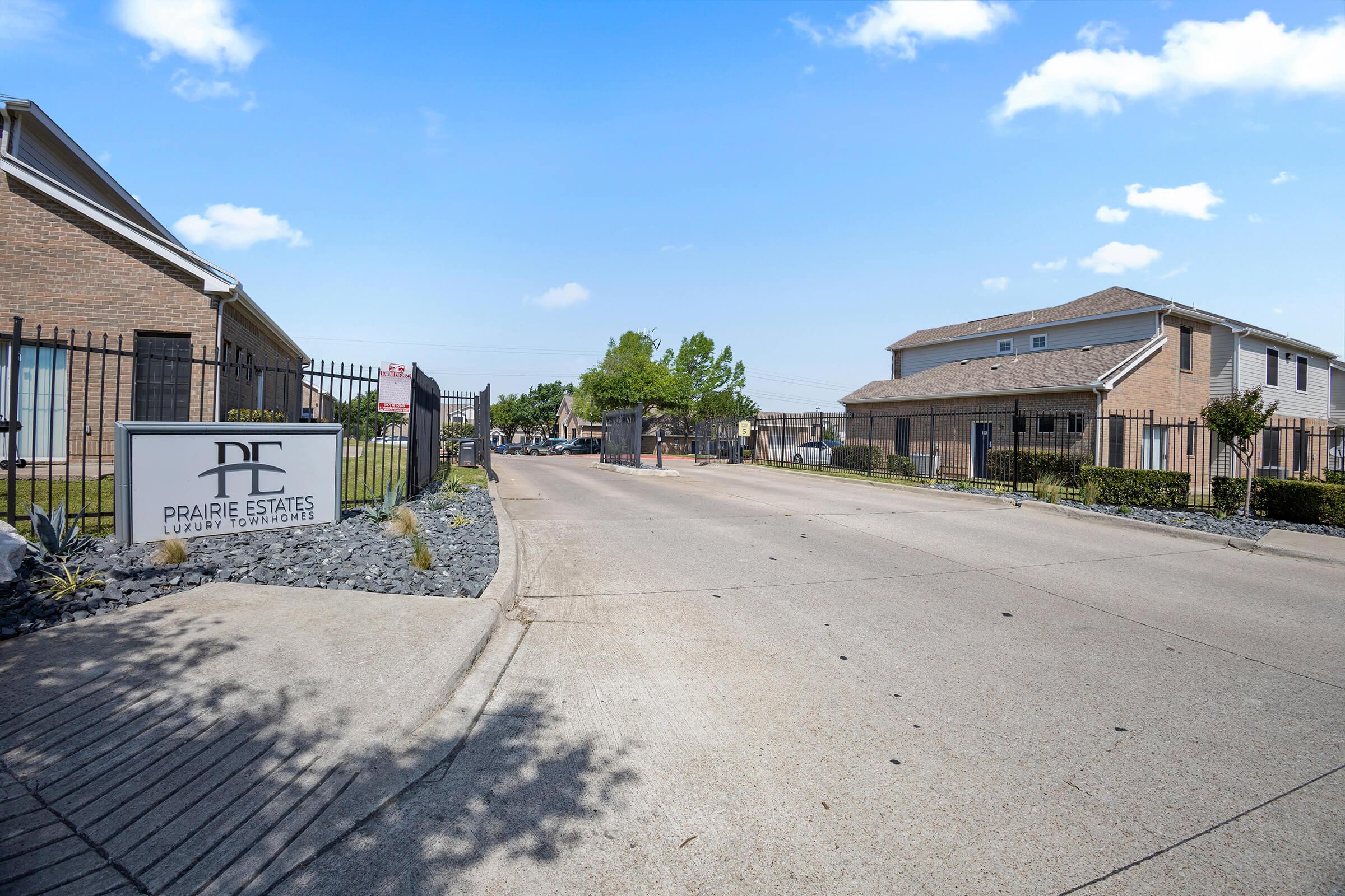 Entrance to Prairie Estates, a residential community. A sign displays the name "Prairie Estates Luxury Apartment Homes," with a gated entrance ahead. On either side, there are well-maintained buildings and landscaped areas, under a clear blue sky. The road ahead is paved and leads into the community.