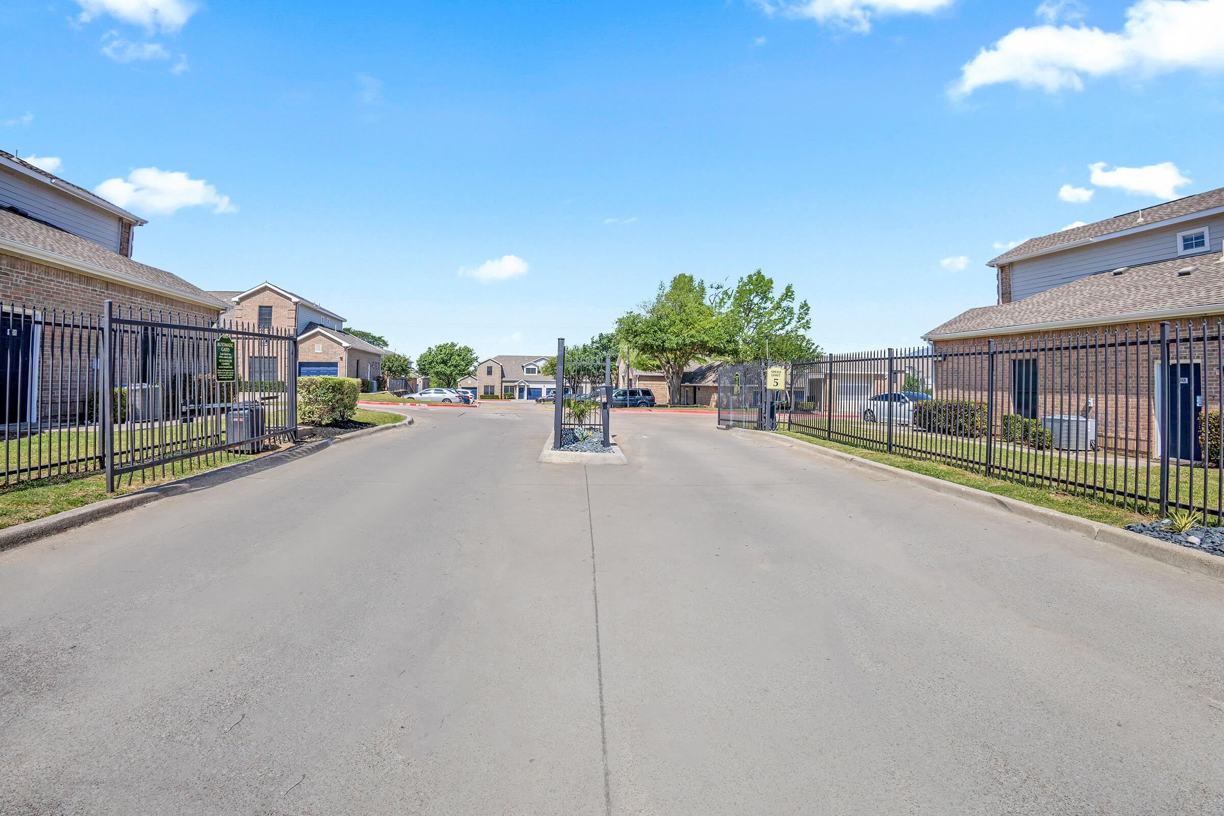 Gated entrance to a residential community, with a paved road leading between two buildings. Lush green lawns and trees are visible on either side, and the sky above is clear with a few scattered clouds. The gate is partially open, inviting entrance into the area.