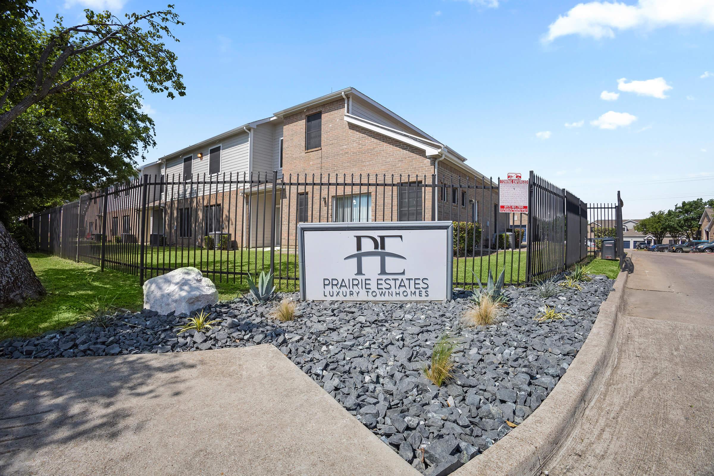 A well-kept sign for Prairie Estates Luxury Townhomes displayed outside a brick building. The sign is surrounded by landscaped rocks and small plants, with a clear blue sky above. The area appears tidy and inviting, indicating a residential community.