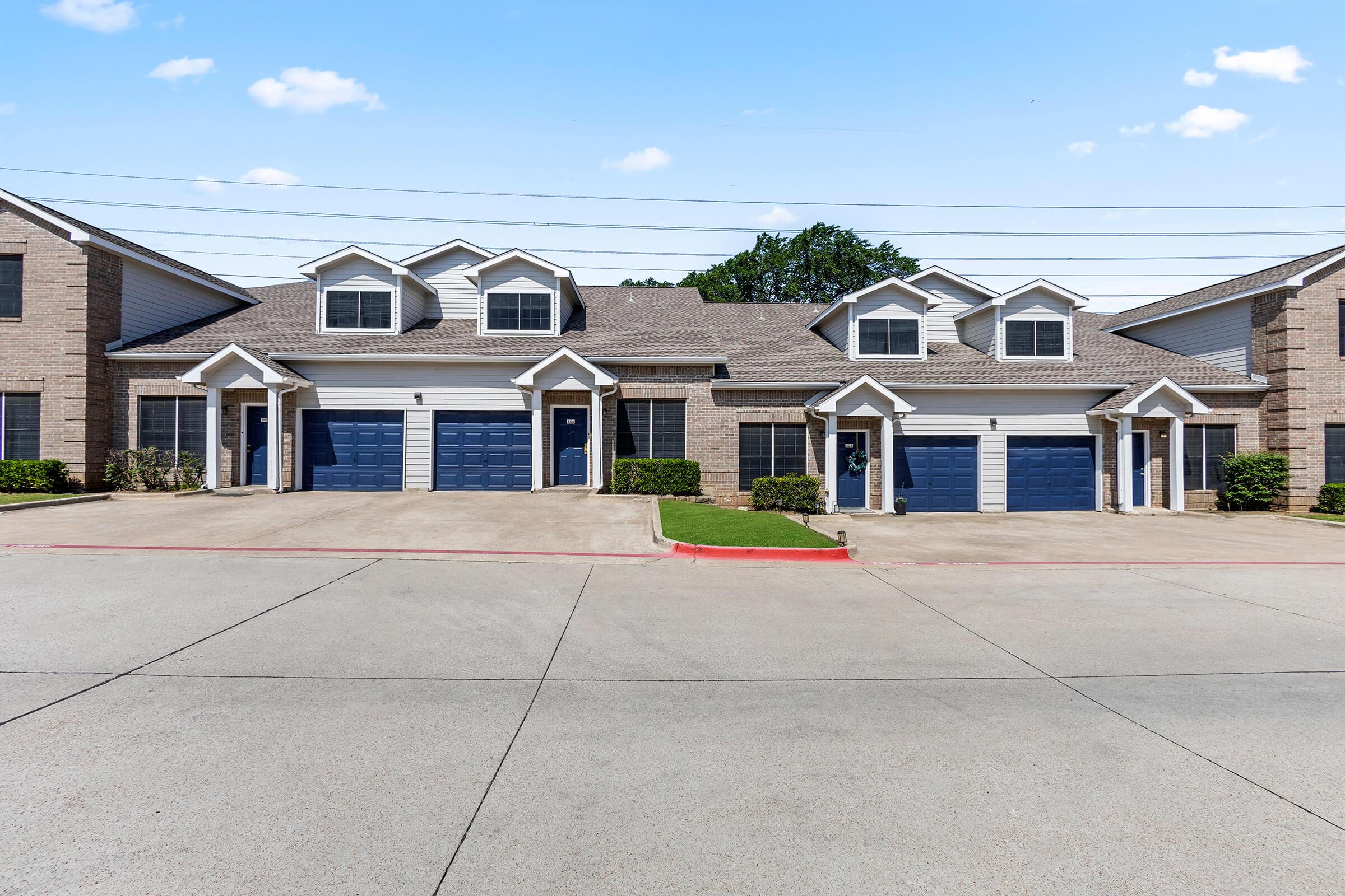 A row of two-story residential townhouses with blue garage doors and well-maintained landscaping. The buildings feature a mix of brick and siding exteriors, and there is a paved driveway leading up to the garages. The sky above is clear with a few clouds.