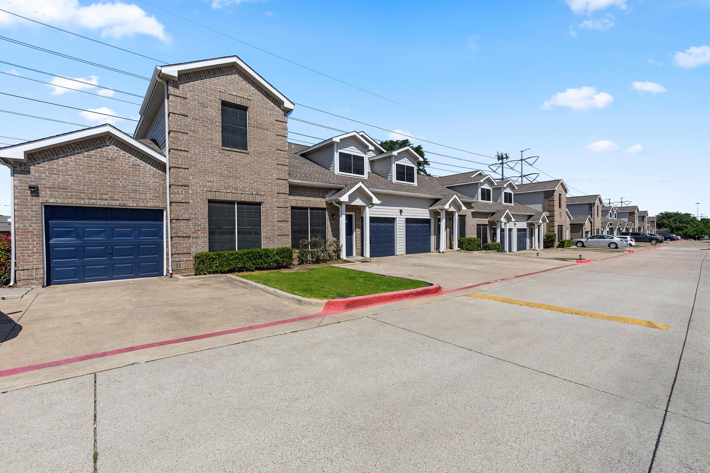 Row of modern brick townhouses with garages, neatly lined on a paved street. The exterior features large windows and a clean landscape, with power lines visible in the background. The sky is clear with a few clouds, suggesting a sunny day.