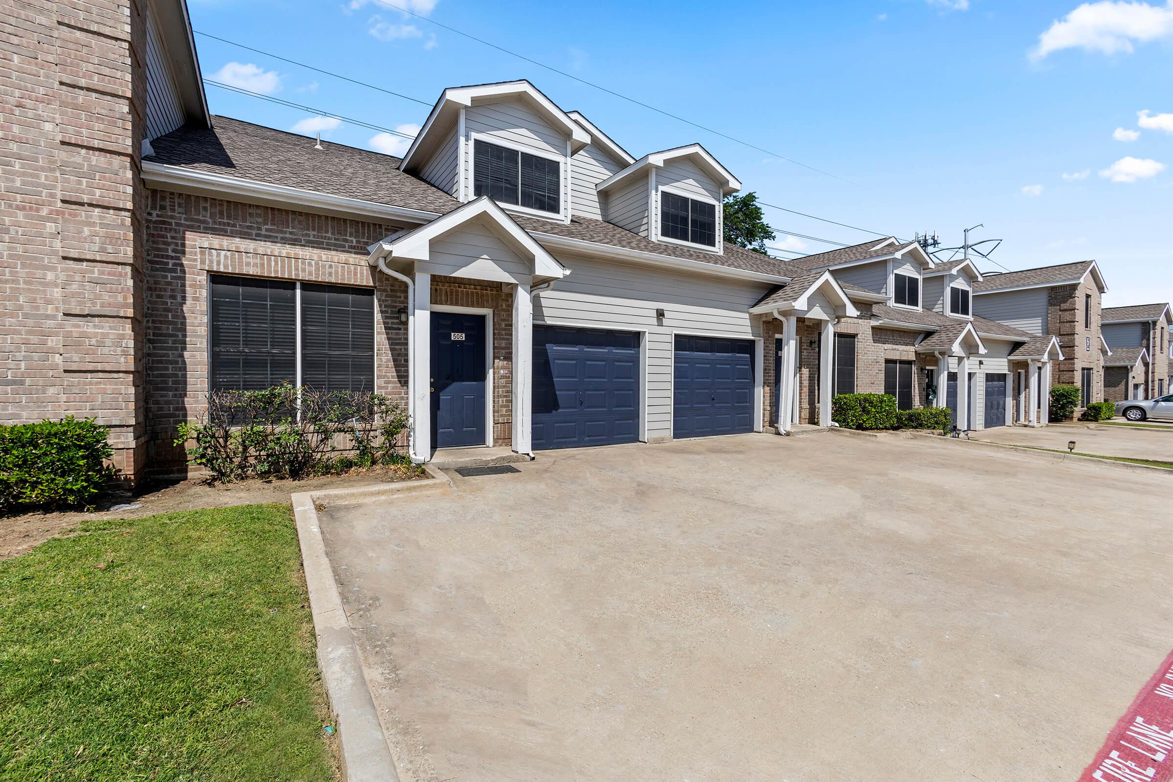 A row of modern townhomes with brick exteriors and dark blue garage doors. Each unit features a small front yard with shrubs, and clear blue skies are visible in the background. The area appears well-maintained, with an empty driveway in the foreground.