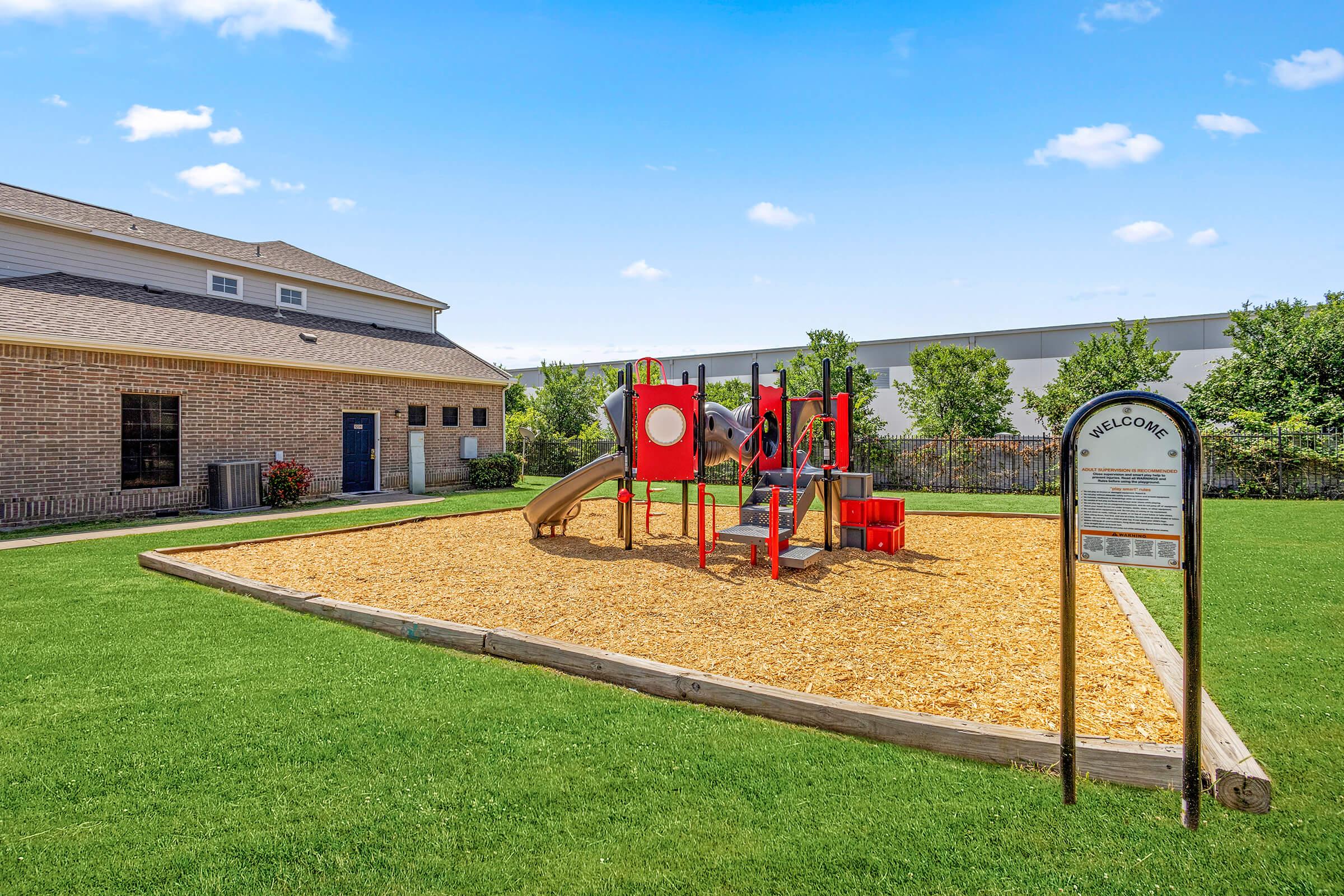 A colorful playground featuring a slide, climbing structures, and safety surfacing made of wood chips, situated on a grassy area. In the background, there is a building with a blue door and landscaping with shrubs. A welcome sign is positioned nearby, adding a community feel.