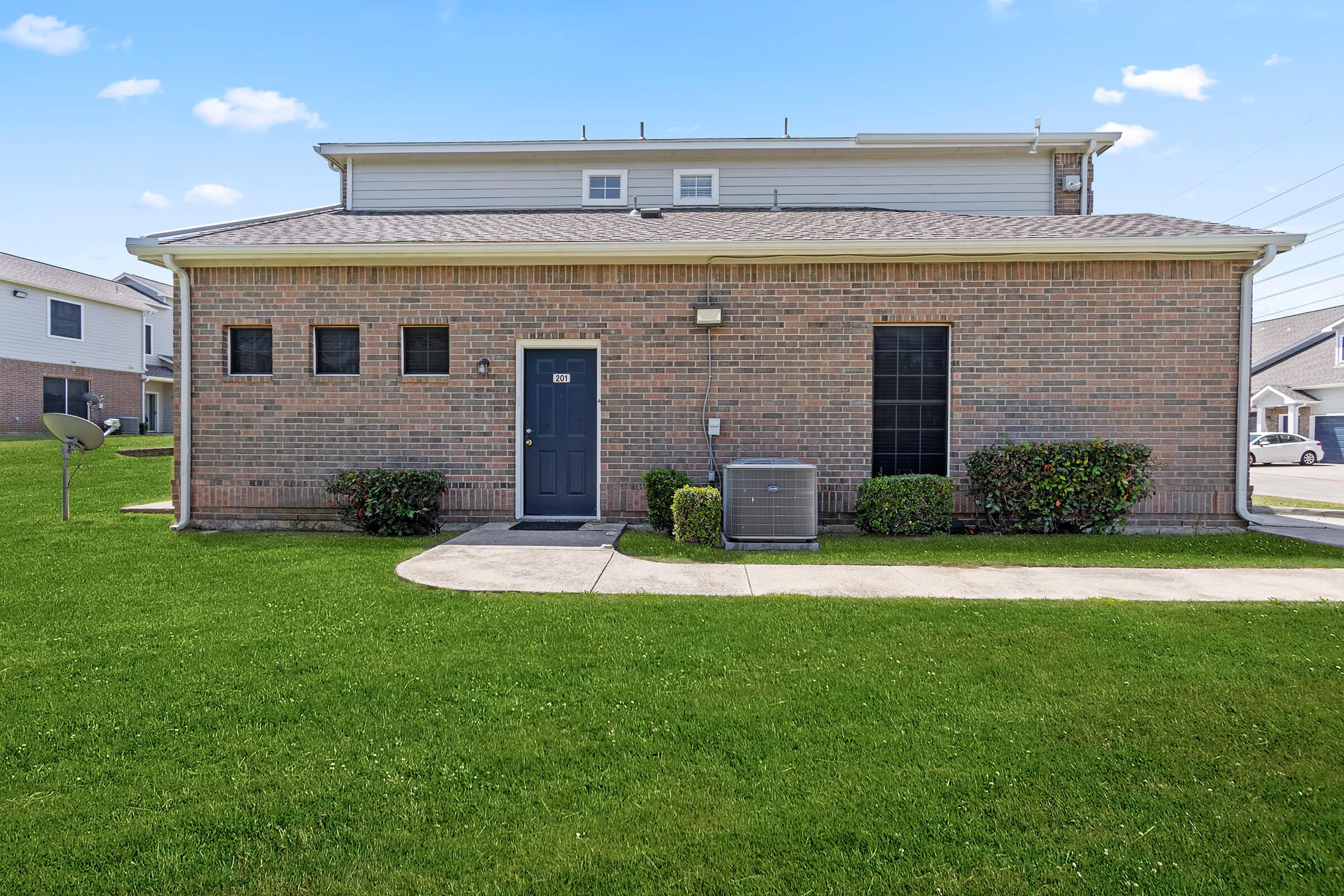 A residential building with a brick exterior, featuring a dark blue door and two windows. The front yard is well-maintained with green grass and bushes. There is an air conditioning unit on the side, and a sidewalk leads up to the entrance. The sky is clear and blue, adding to the pleasant appearance of the property.