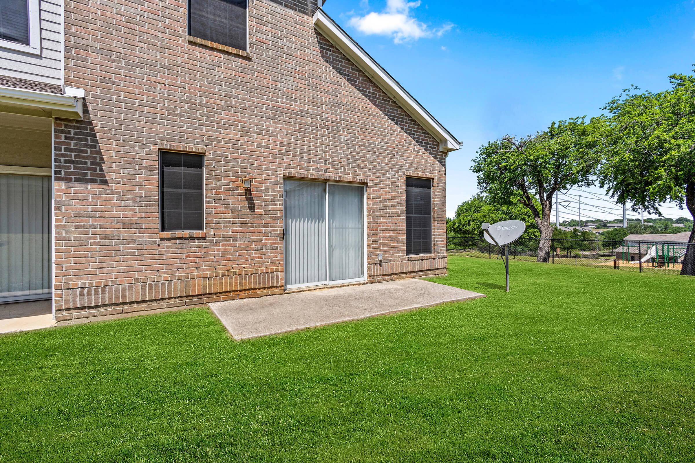 Exterior view of a house featuring a brick wall, a sliding glass door, and a patio area. A satellite dish is installed on the grassy lawn, with trees in the background and a clear blue sky.