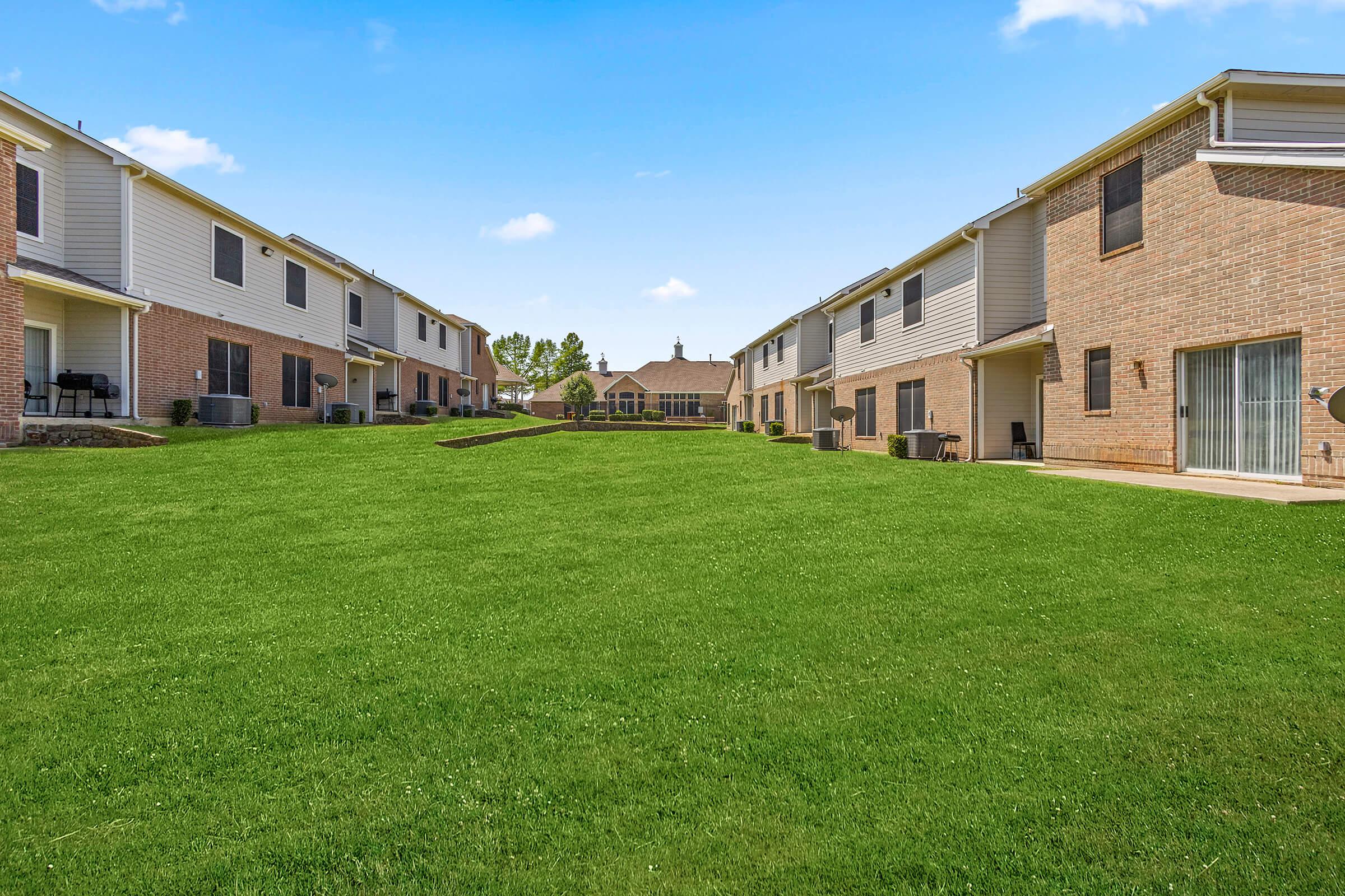 A landscaped green space between two rows of residential buildings, featuring well-maintained grass and a clear blue sky. In the background, a community building is visible, surrounded by trees. The scene conveys a peaceful residential atmosphere.
