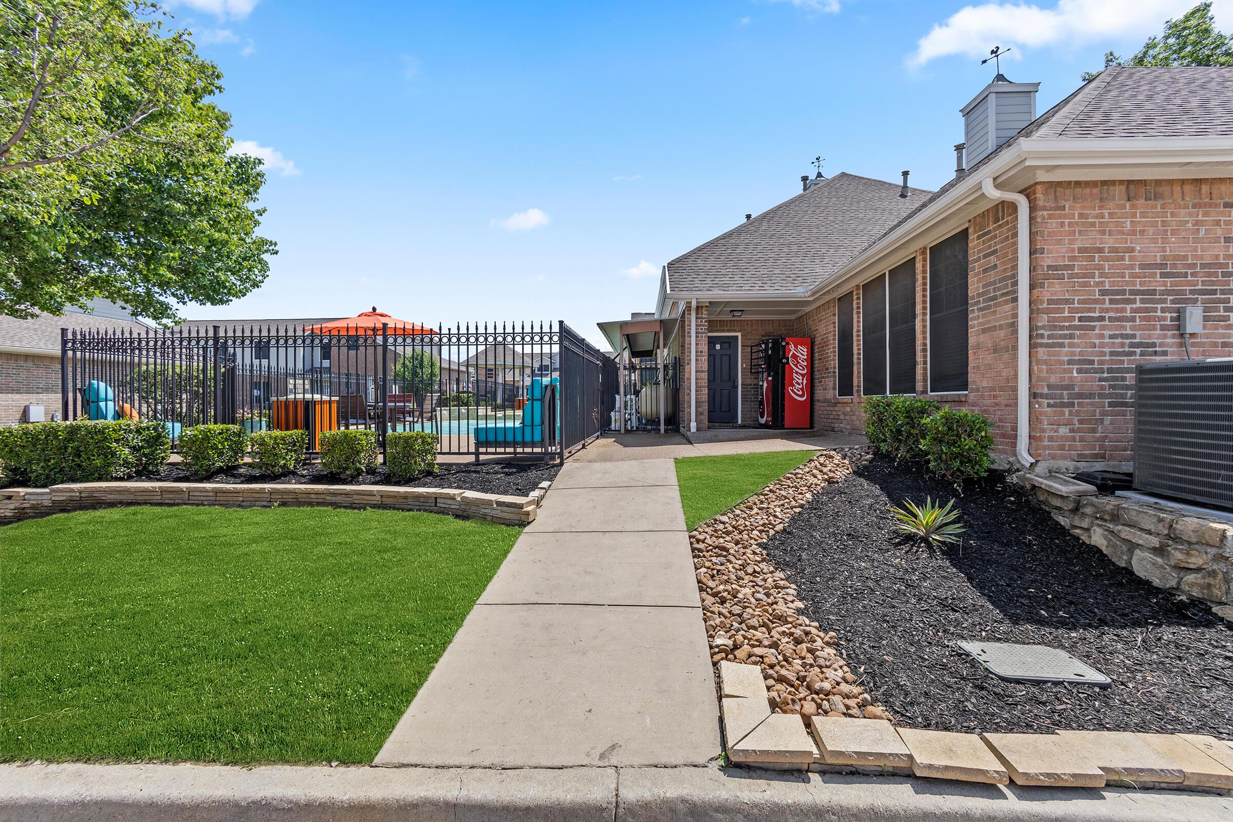 A well-maintained pathway leads to a brick building with a Coca-Cola vending machine outside. Lush green grass and decorative stones line the path, while a fenced area in the background features colorful outdoor furniture and a pool. The scene is bright and inviting under a clear blue sky.
