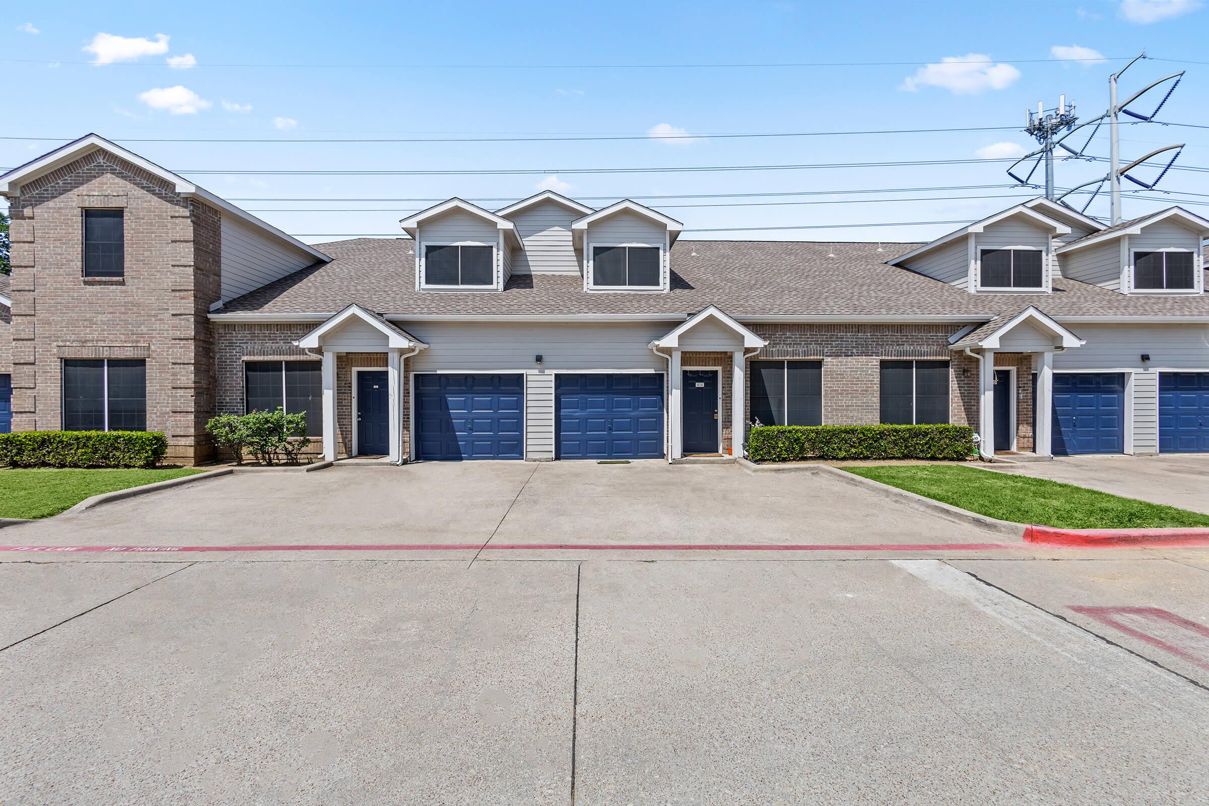 Two-story residential building with multiple garage doors, featuring brick and siding design. The property has green shrubbery in front and a clear blue sky above. Power lines are visible in the background, and the driveway is paved, indicating a well-maintained neighborhood.