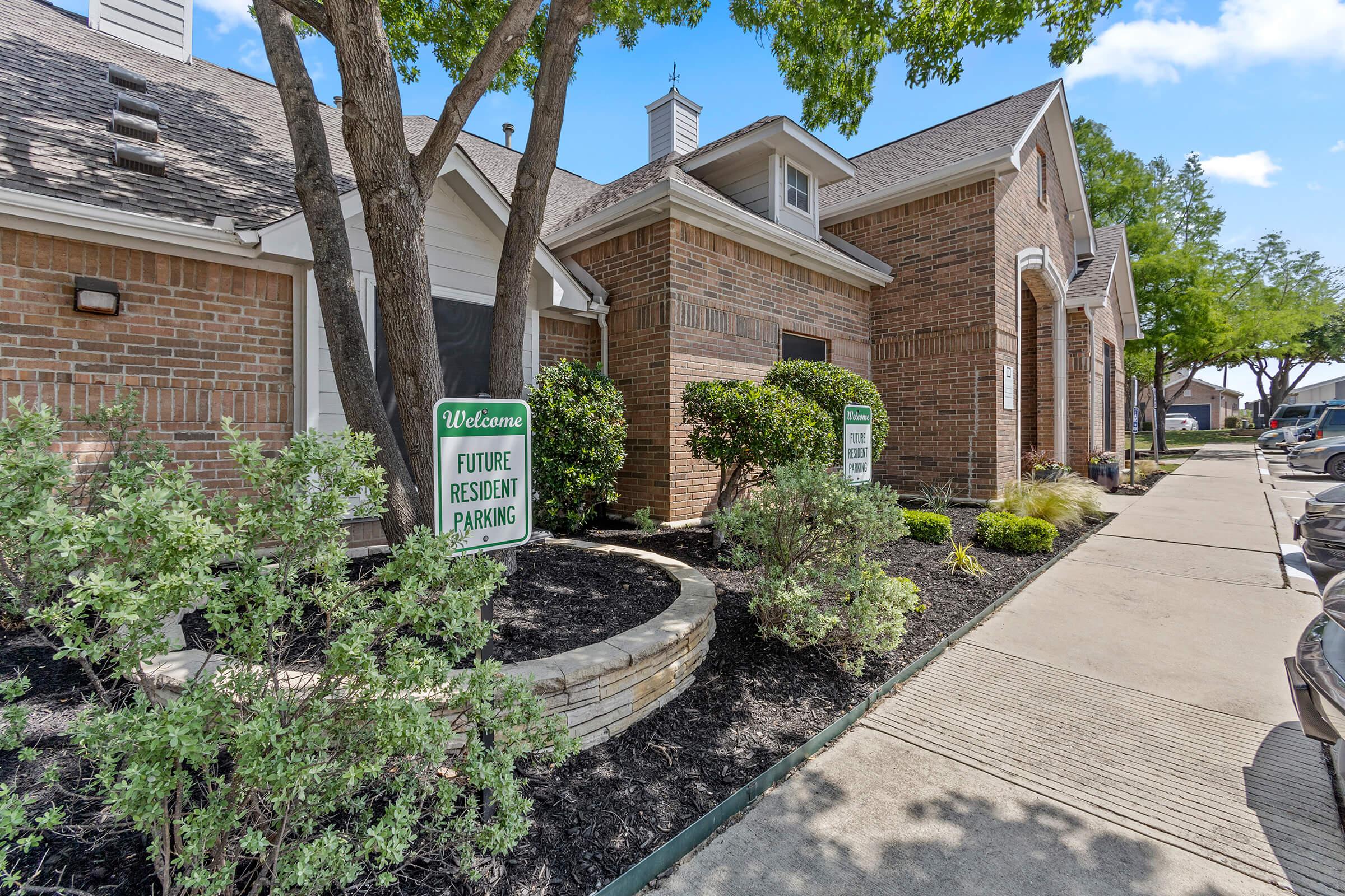 A well-maintained residential complex showcasing a brick building with landscaped greenery. Signs indicate "Welcome" and "Future Resident Parking." The pathway is lined with bushes and trees, enhancing the inviting atmosphere of the entrance area. Clear blue skies complement the scene.