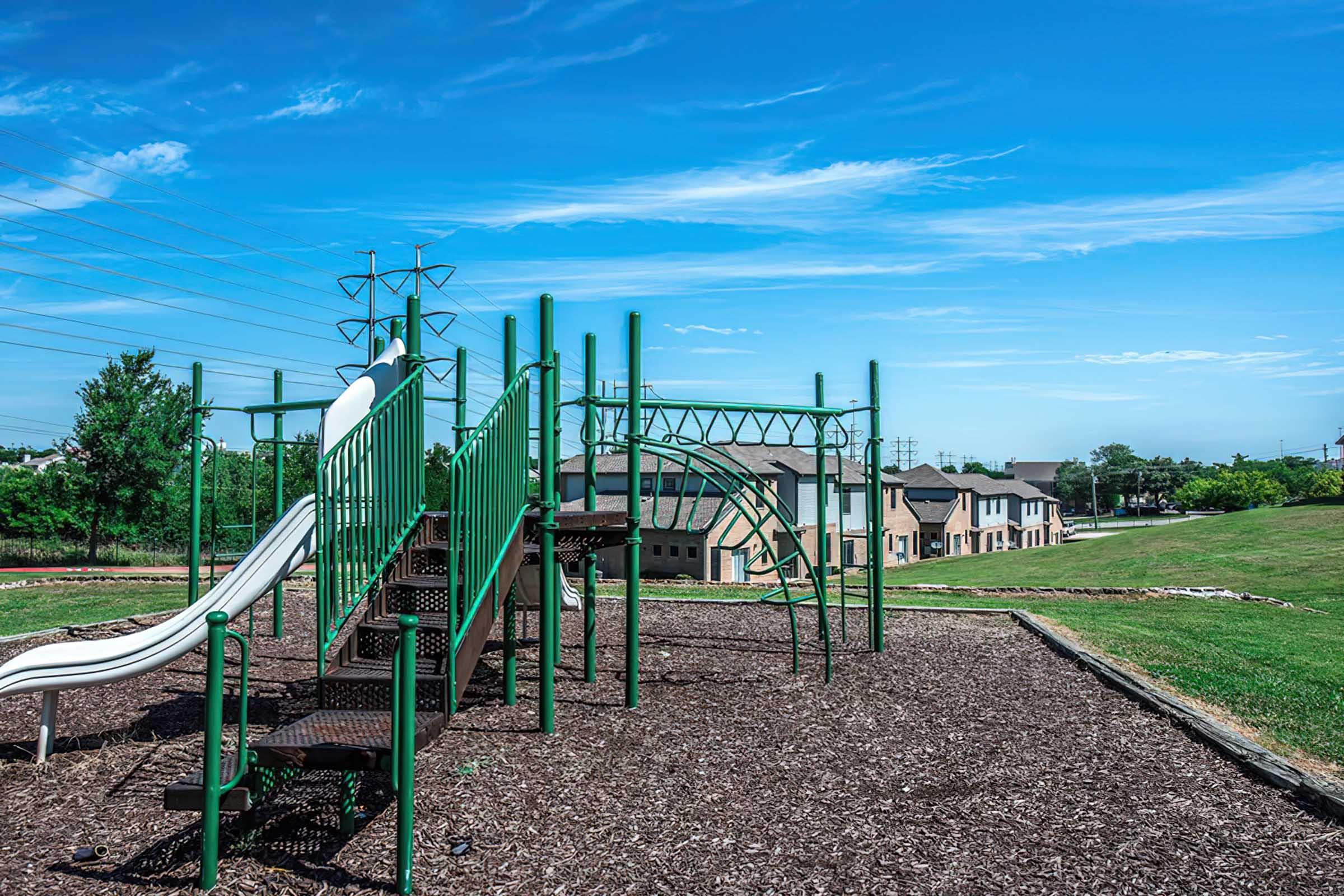 A playground featuring a green slide and climbing structure, surrounded by mulch. In the background, there are residential buildings under a clear blue sky with a few clouds and power lines. The scene conveys a bright, sunny day with ample green space.