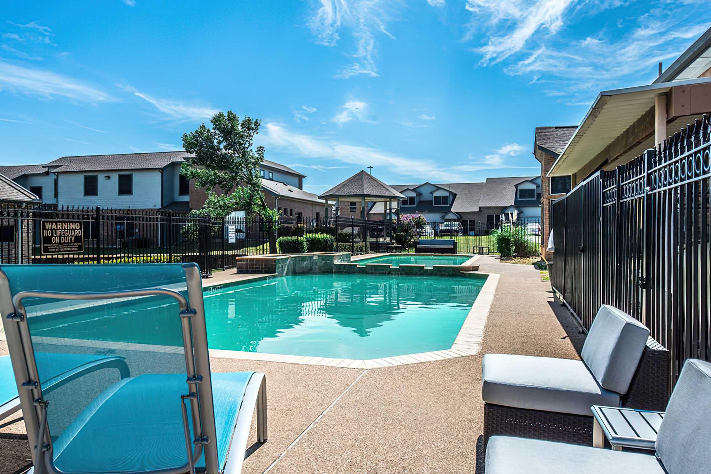 A clear blue swimming pool surrounded by a concrete patio, with lounge chairs nearby. In the background, green grass and landscaped areas are visible, along with residential buildings and a gazebo under a bright blue sky. A warning sign is posted near the pool area.
