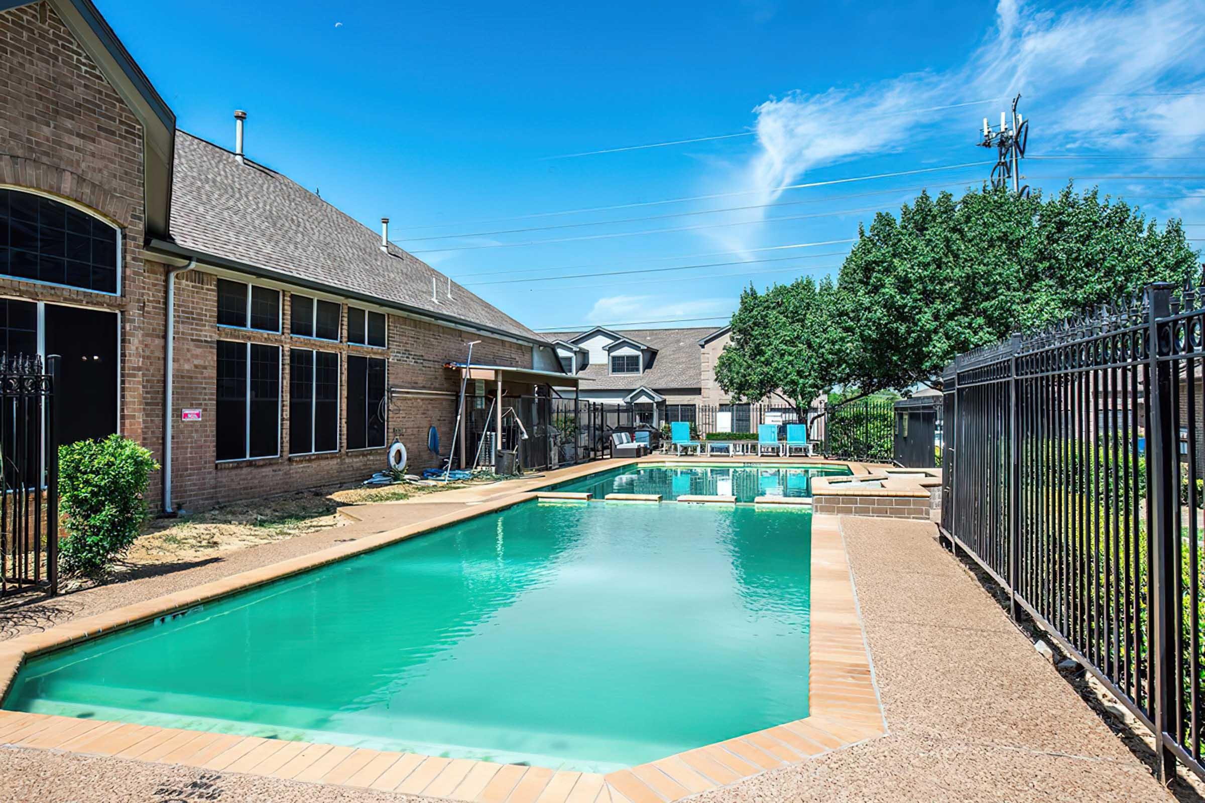 A tranquil swimming pool area featuring two pools with clear blue water, surrounded by a wrought iron fence. Lush green trees provide shade, and a building with large windows is visible in the background under a clear blue sky. The scene conveys a serene atmosphere ideal for relaxation.
