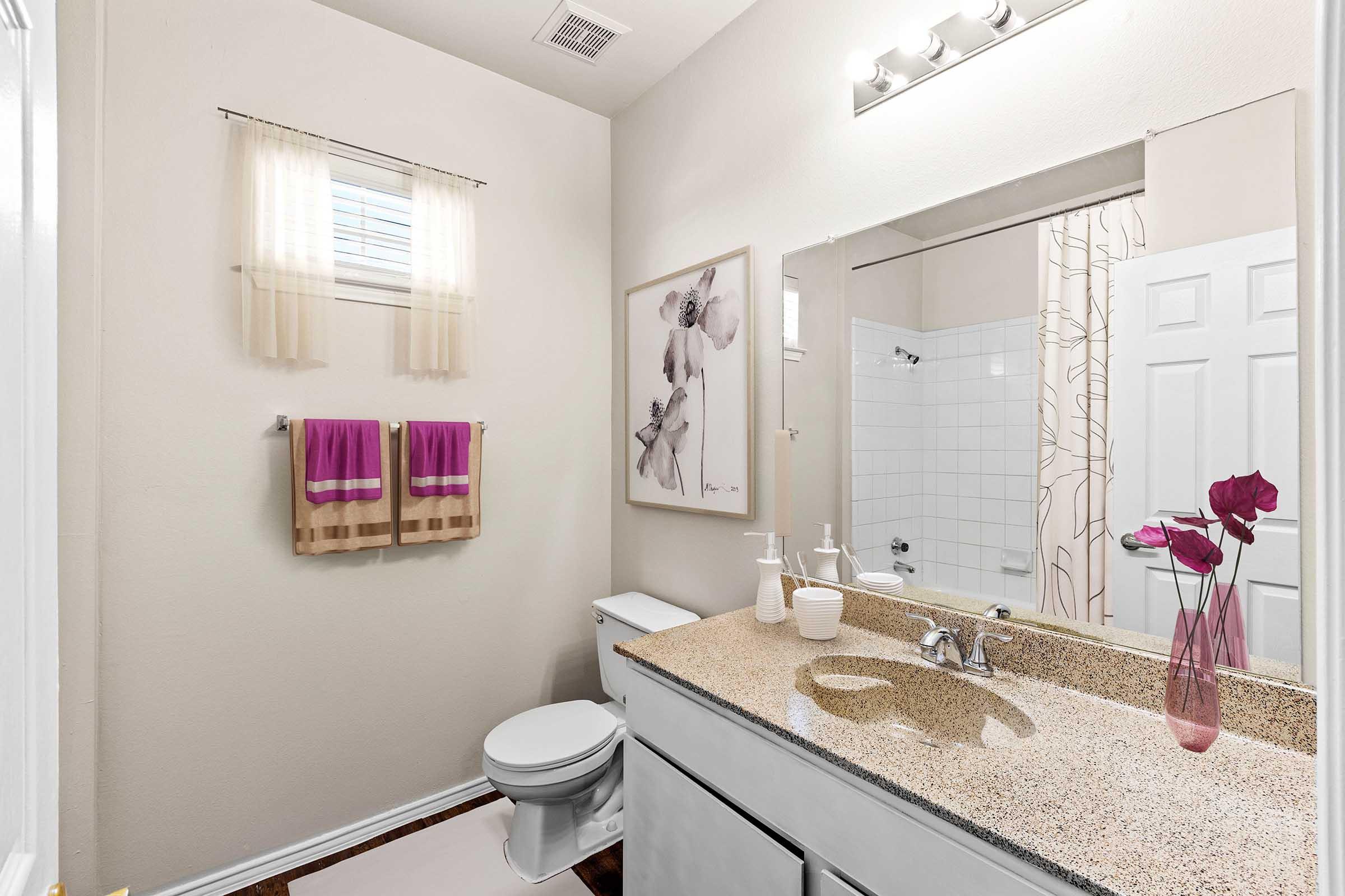 A clean and modern bathroom featuring a white toilet, a sink with a granite countertop, and a large mirror. Two purple towels hang on a rack, and a framed floral artwork is displayed on the wall. There is a shower area with curtains, and a small vase with flowers on the counter. Natural light comes in through a window.