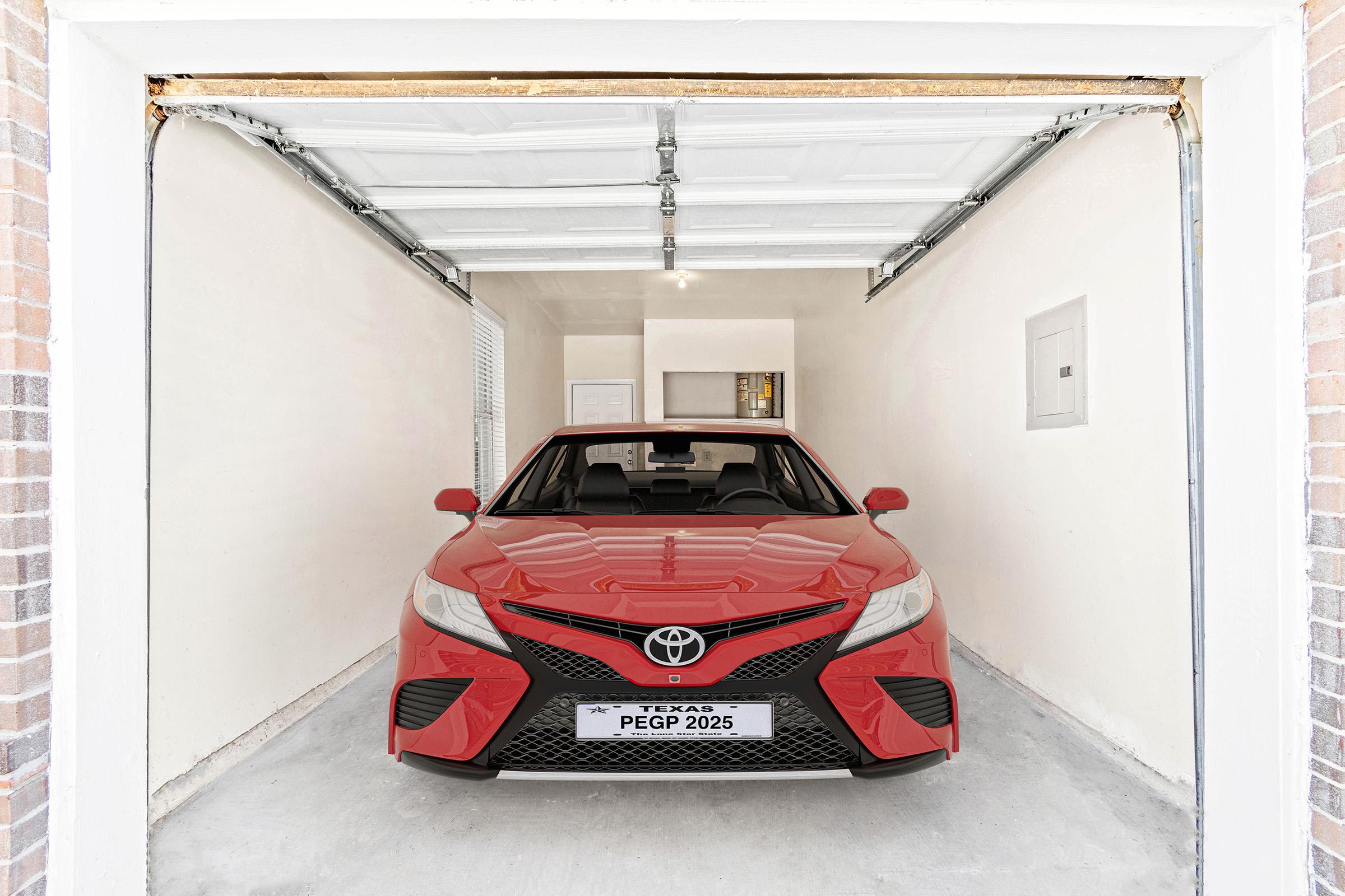 A red Toyota Camry is parked in a clean, well-lit garage with a white interior. The garage door is open, allowing natural light to enter. A license plate with "TEXAS PEGP 2025" is visible on the front of the car. The overall space appears neat and organized.