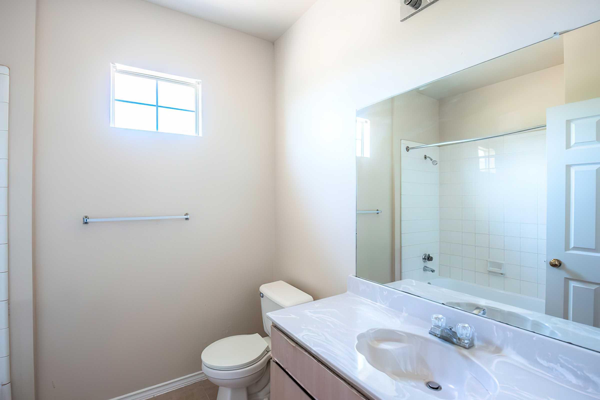 A clean, well-lit bathroom featuring a white tiled shower, a toilet, and a double sink vanity with a large mirror. There's a window allowing natural light to enter, and the walls are painted in a light color, providing a spacious feel. A towel rack is mounted on the wall beside the mirror.