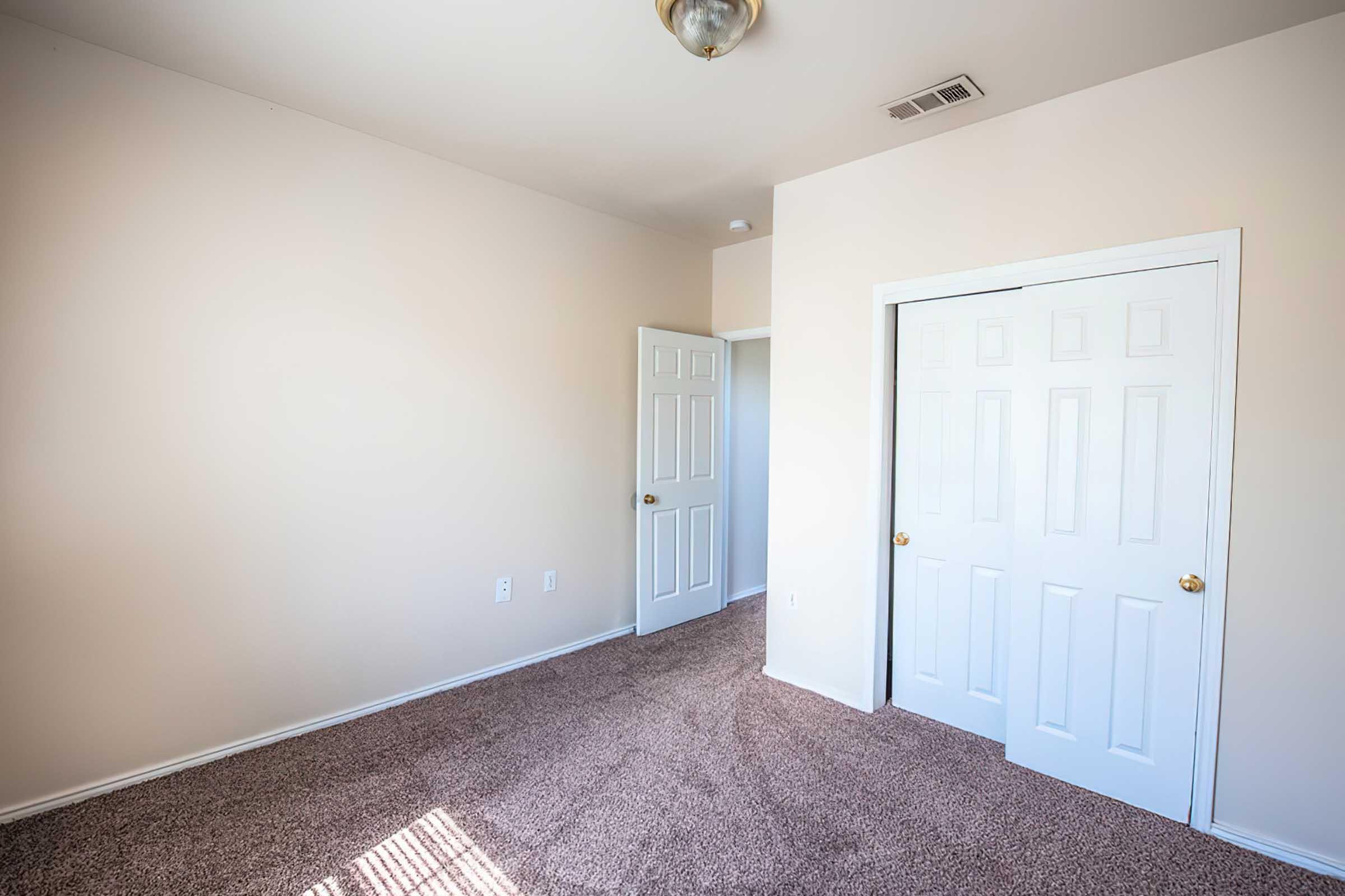 Interior of a small, empty room featuring light beige walls, a carpeted floor, and a ceiling light fixture. There are two white doors: one leading to another room and another closet door. Natural light is coming in, creating soft shadows on the floor.