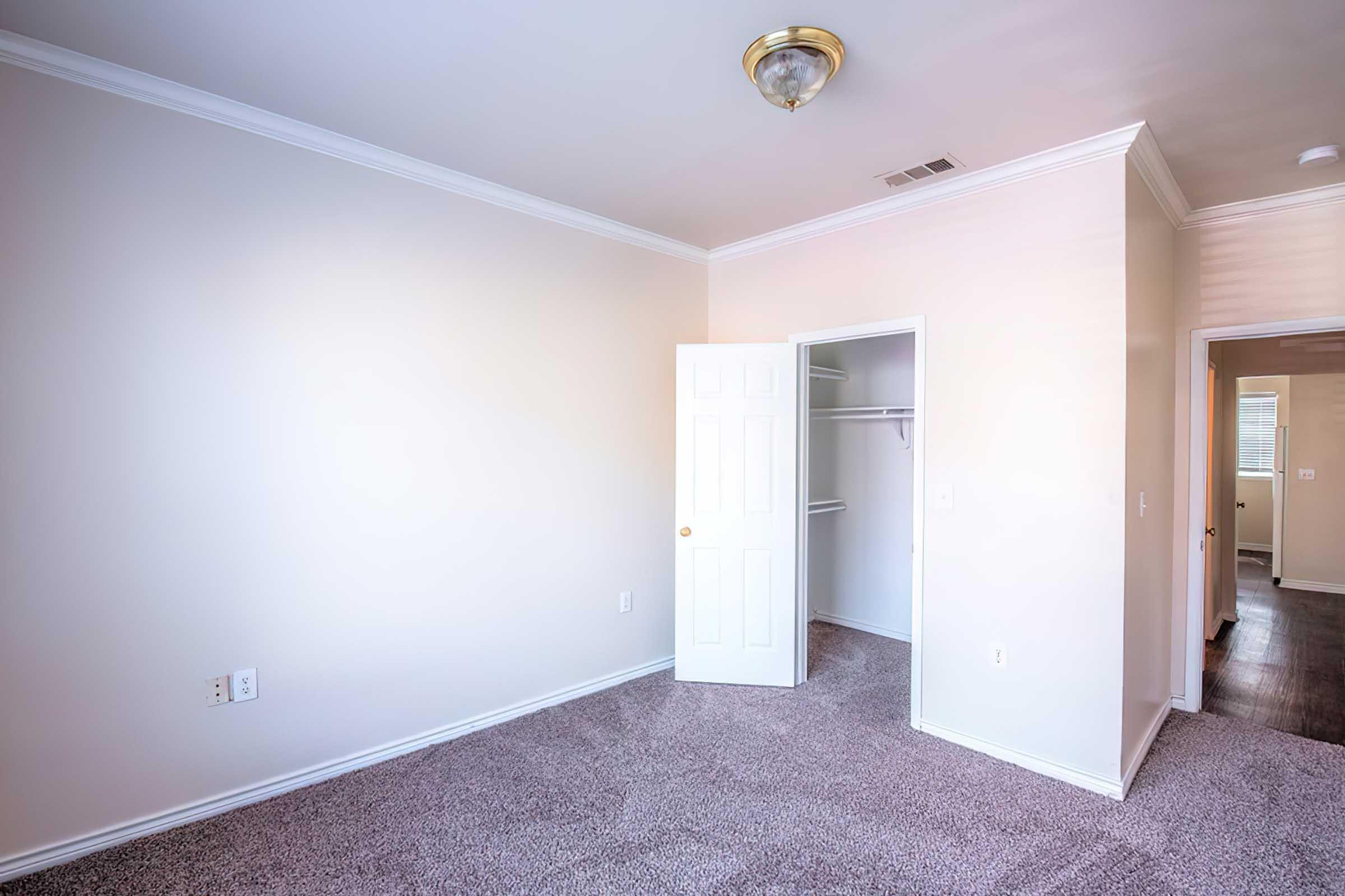 An empty, well-lit bedroom with light-colored walls and carpeted flooring. A white door leads to a closet, and another door opens to an adjacent room. The ceiling features a simple light fixture, creating a bright and spacious atmosphere.