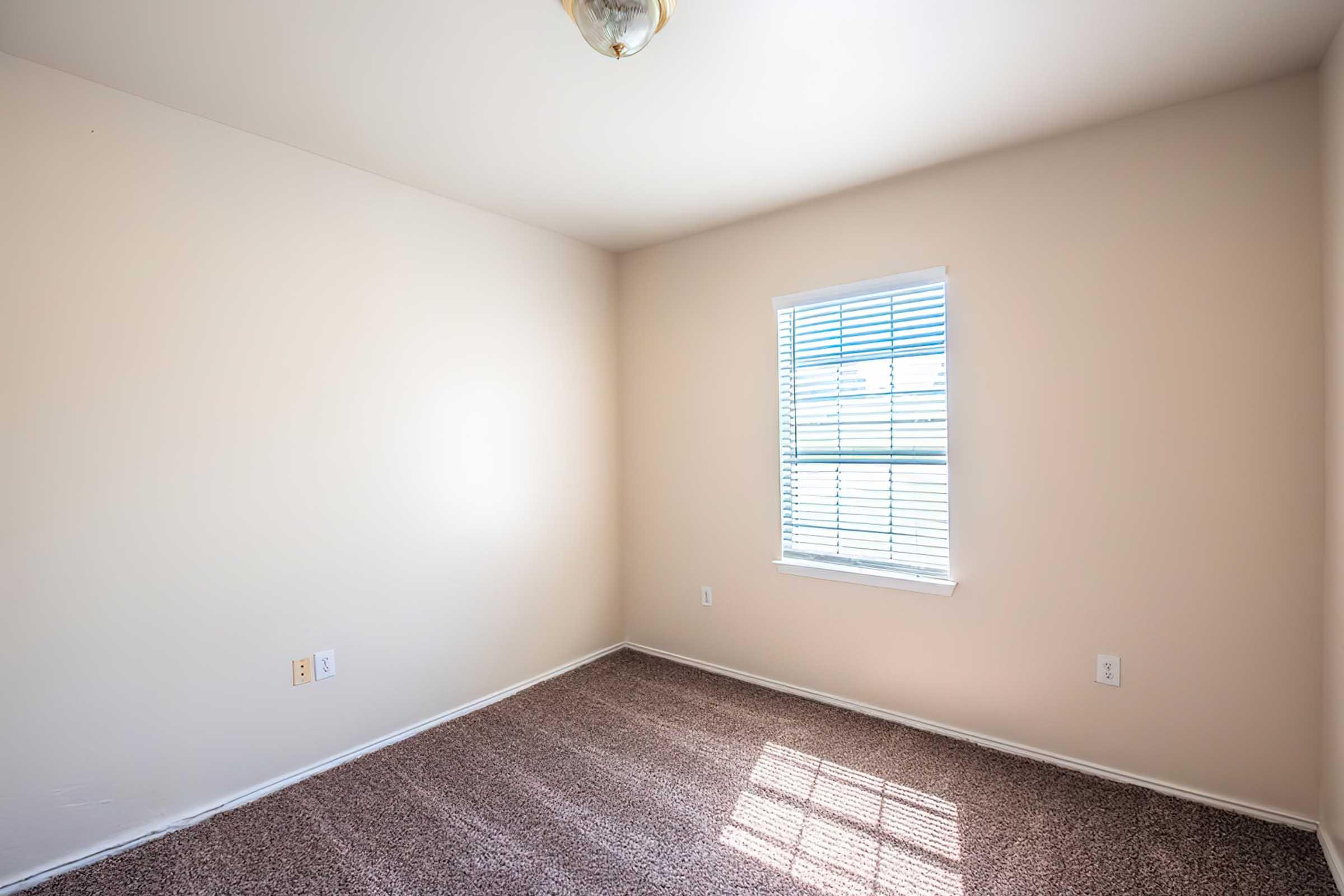 A vacant, empty room with light beige walls and a window featuring white blinds. The floor is covered with brown carpet, and there is a ceiling light fixture. Natural light is coming in through the window, creating a bright atmosphere.
