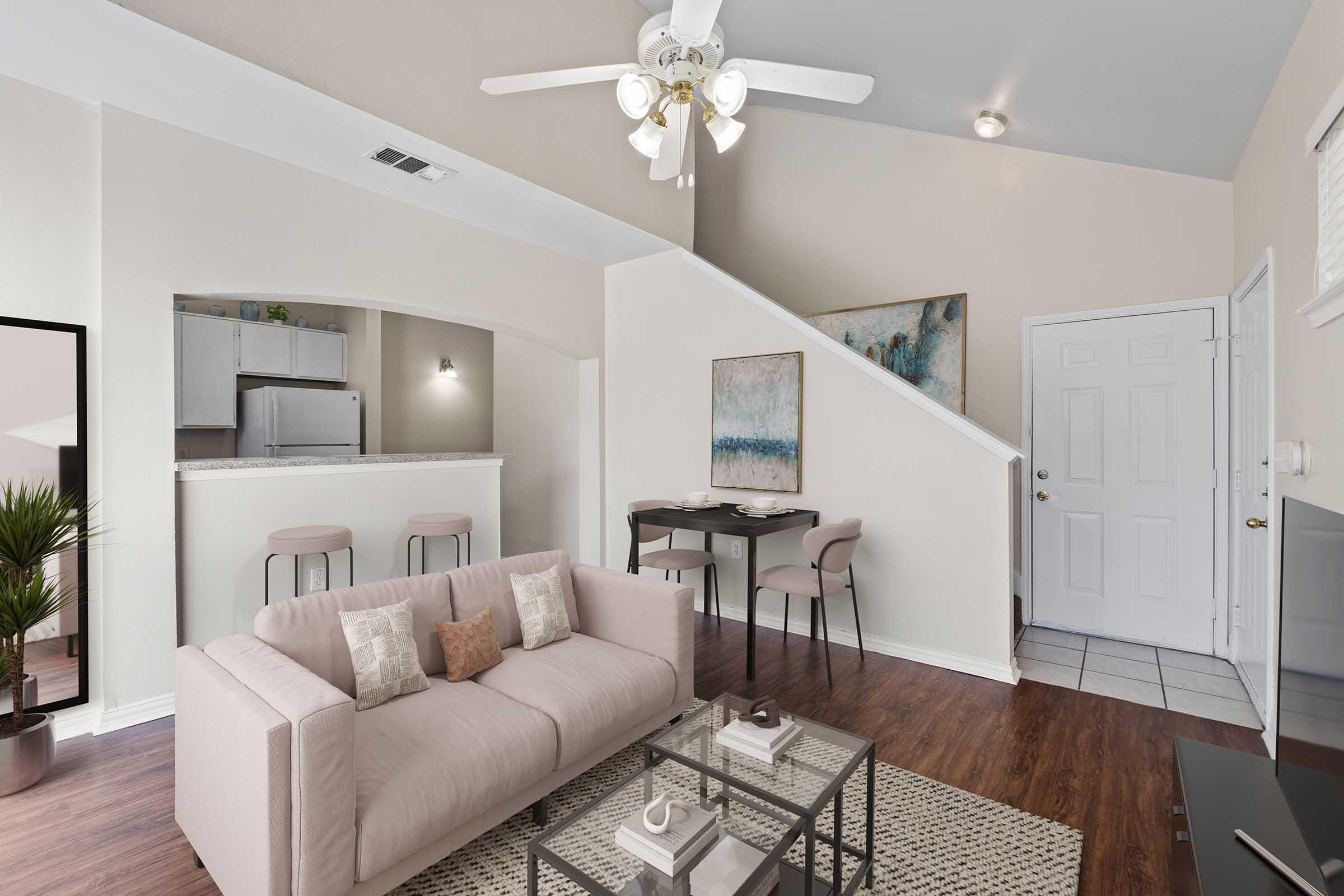 A cozy living room featuring a light pink sofa, a coffee table with decor, and a ceiling fan. There's a dining area with two bar stools and a small table. The open space leads to a kitchen with modern appliances. Light-colored walls and flooring create a warm, inviting atmosphere.