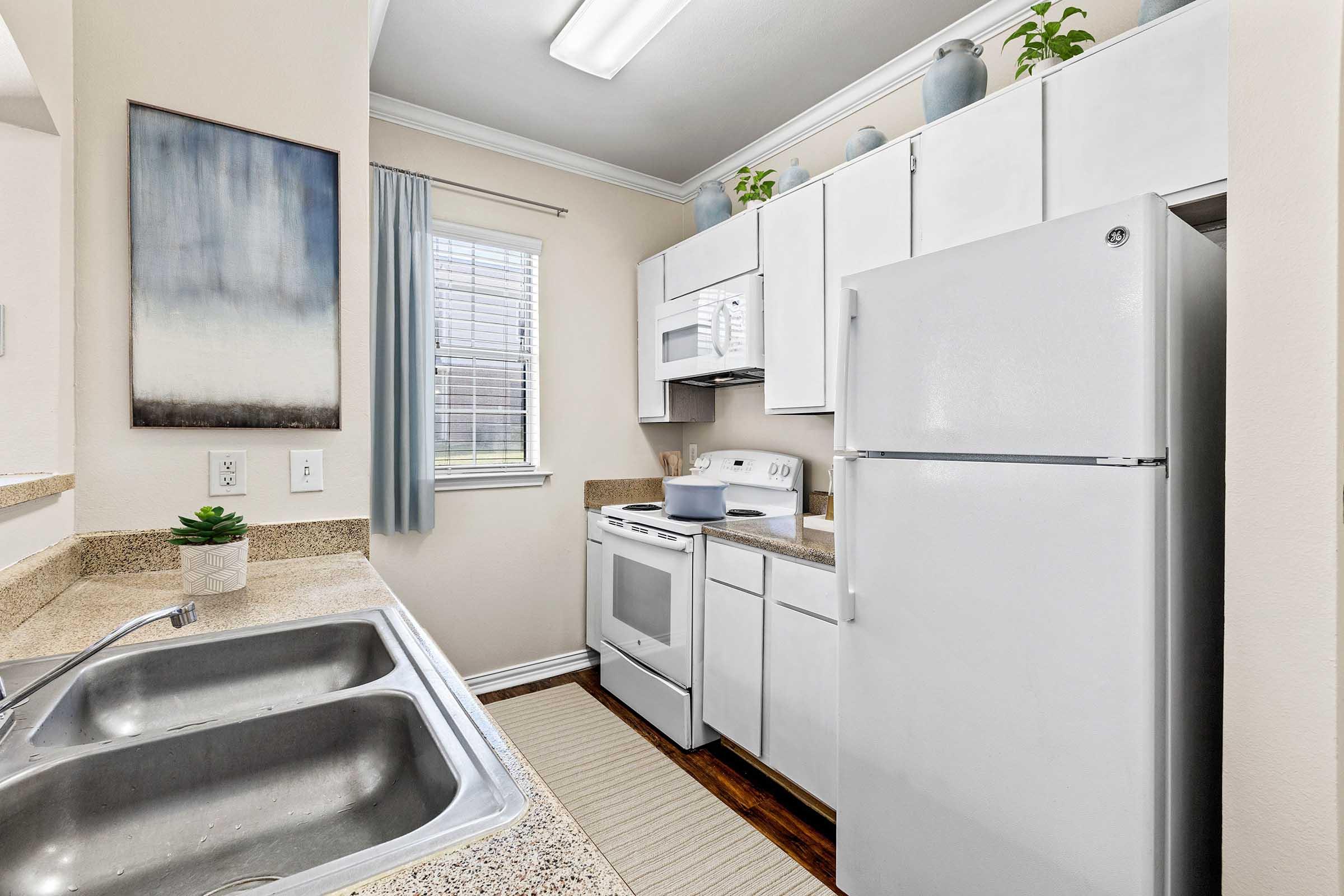 A modern kitchen featuring white cabinets, a double sink, and essential appliances including a refrigerator, microwave, and stove. The countertops are a light color, complemented by subtle decor like potted plants and a piece of wall art. Natural light enters through a window with light curtains.